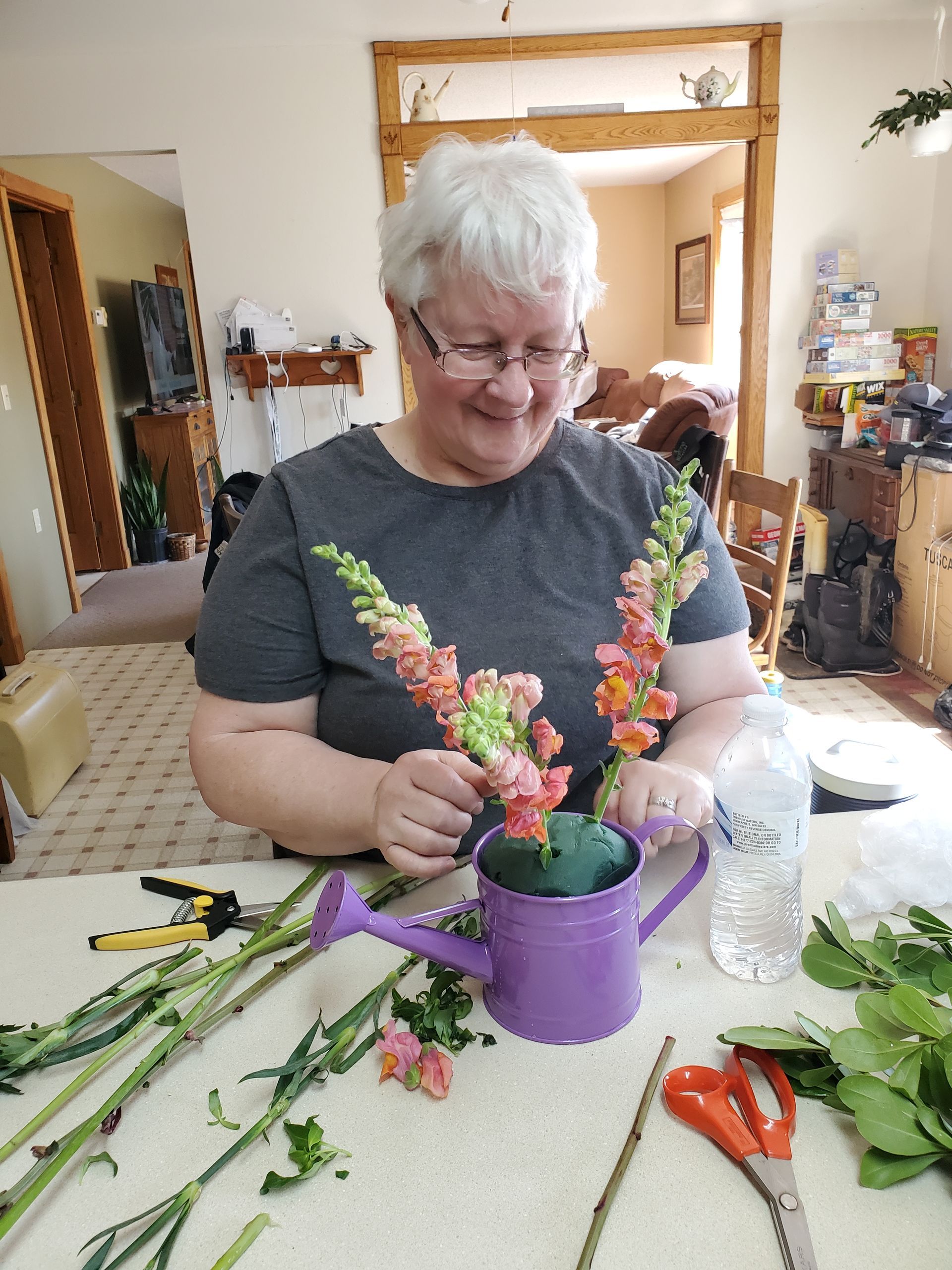 Woman arranging flowers in a purple watering can, smiling. Flowers, clippers, and water are on the table.