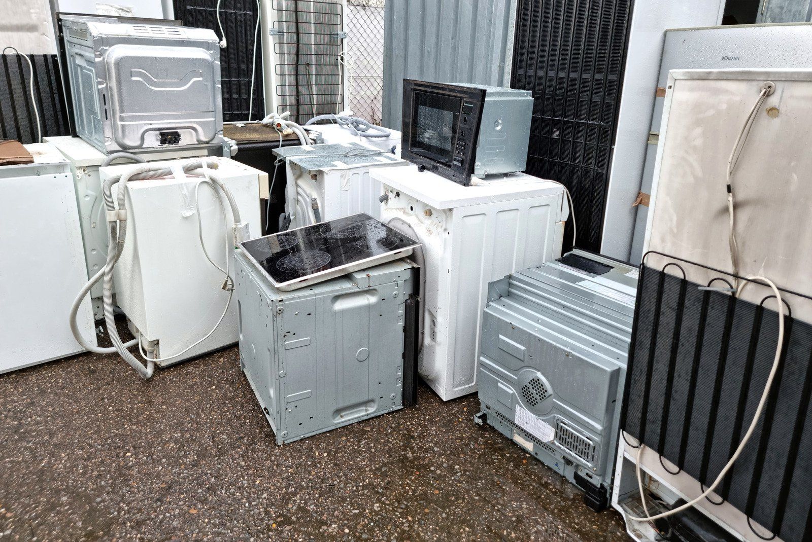 Discarded appliances, including washers, dryer, microwave, and dishwasher, piled on the ground outside.