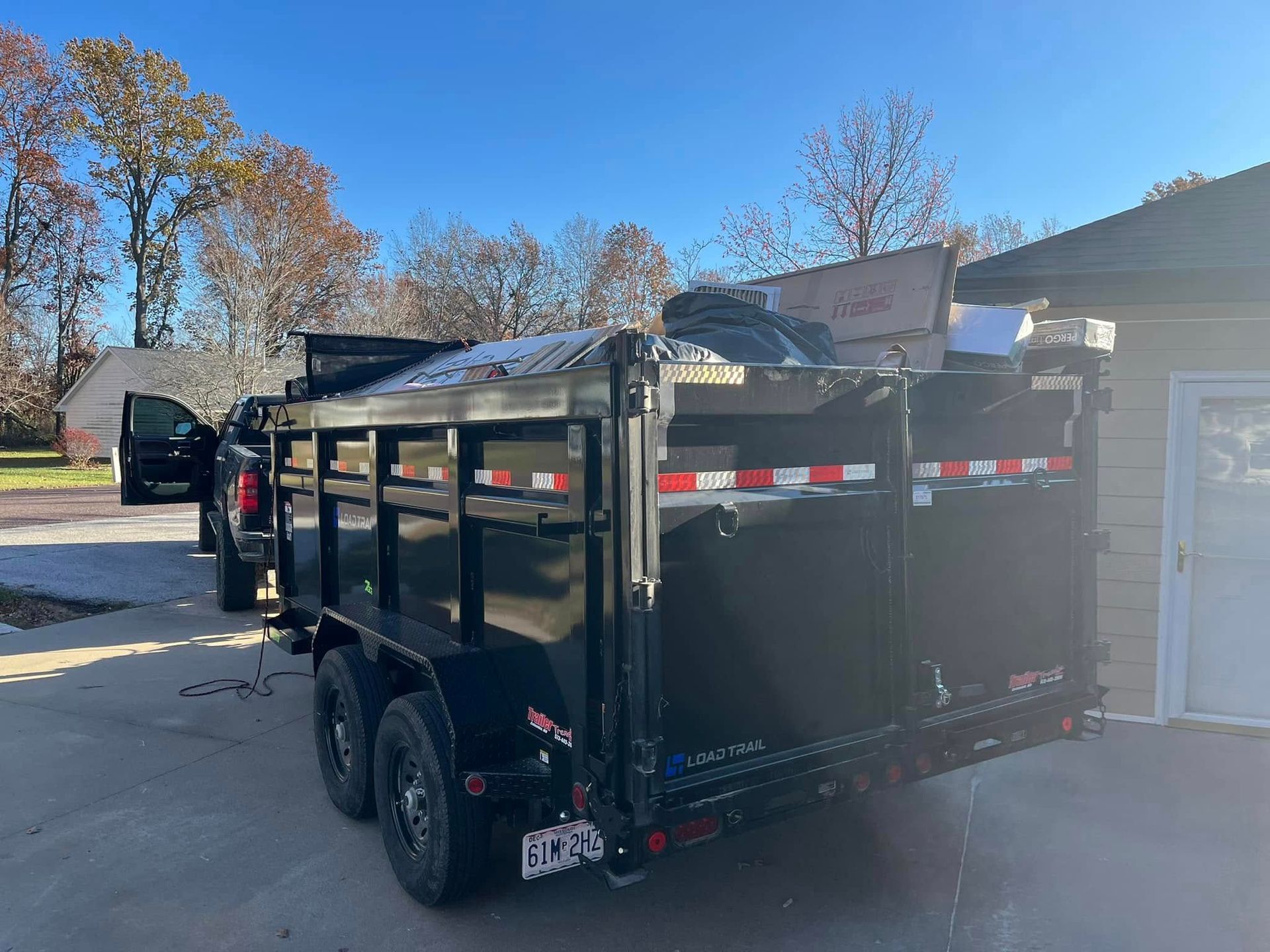 Black dump trailer loaded with debris, hitched to a truck, parked on a driveway in front of a house.