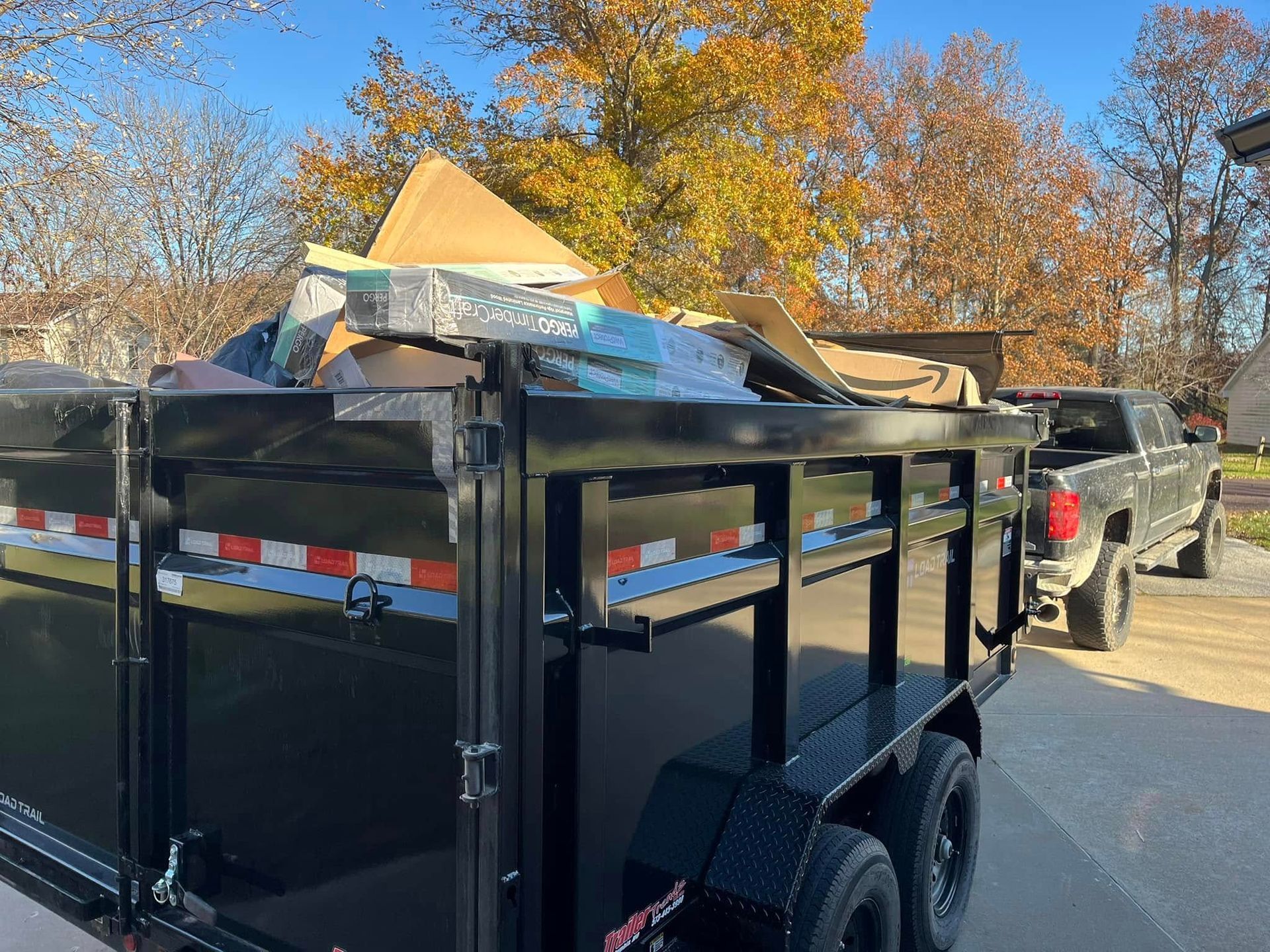 Black trailer loaded with debris, hitched to a truck, set outdoors with trees in the background.