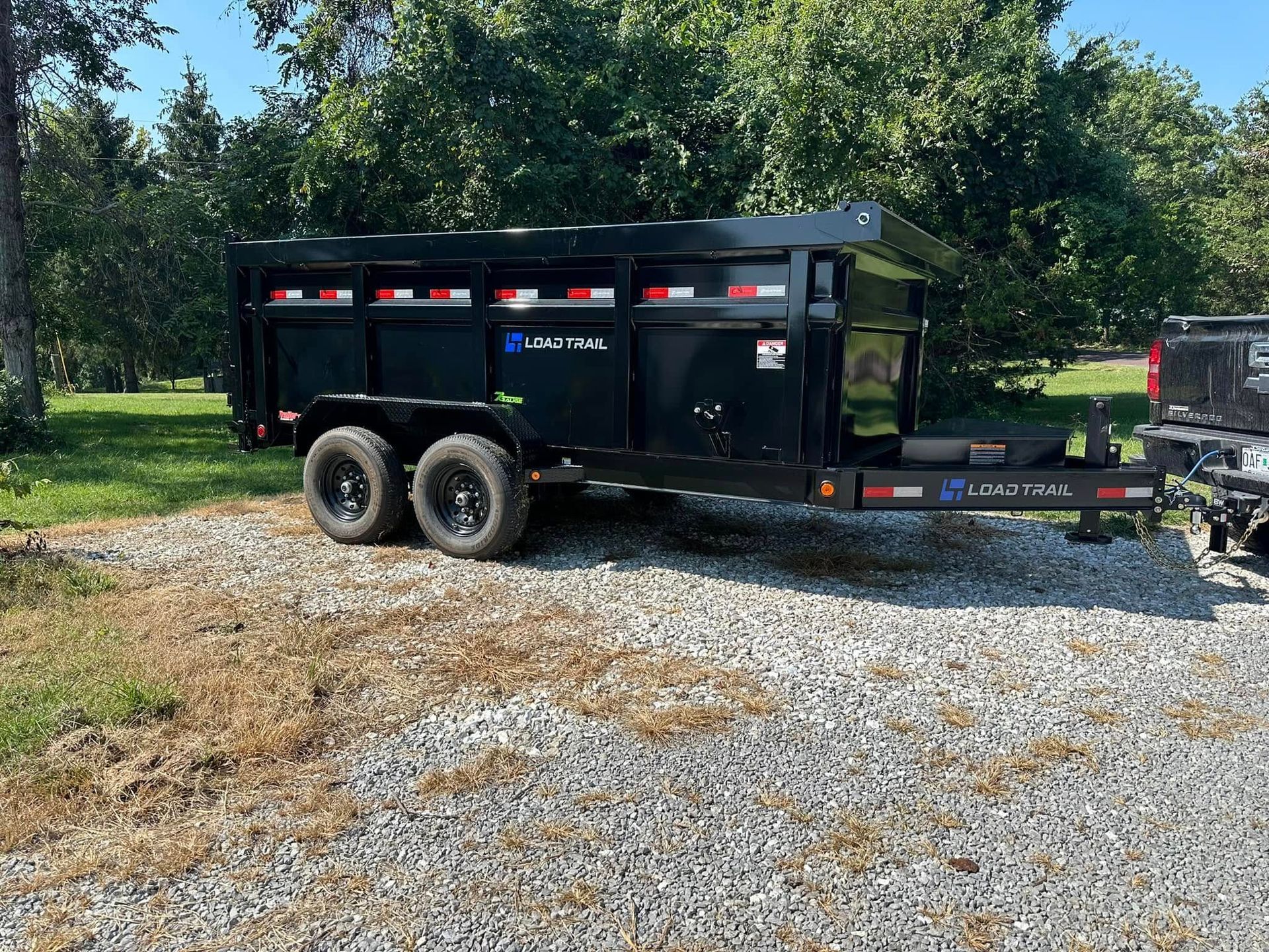 Black dump trailer parked on gravel, with trees in the background.