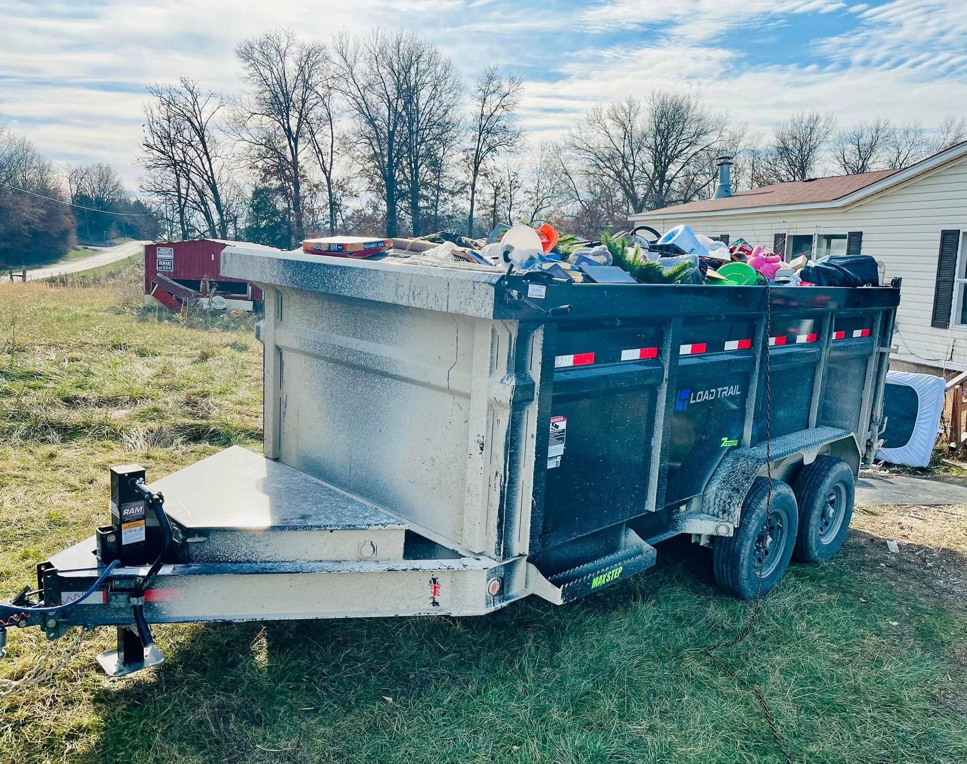 Dump trailer filled with trash, parked in grassy field near a house, trees in the background, overcast day.