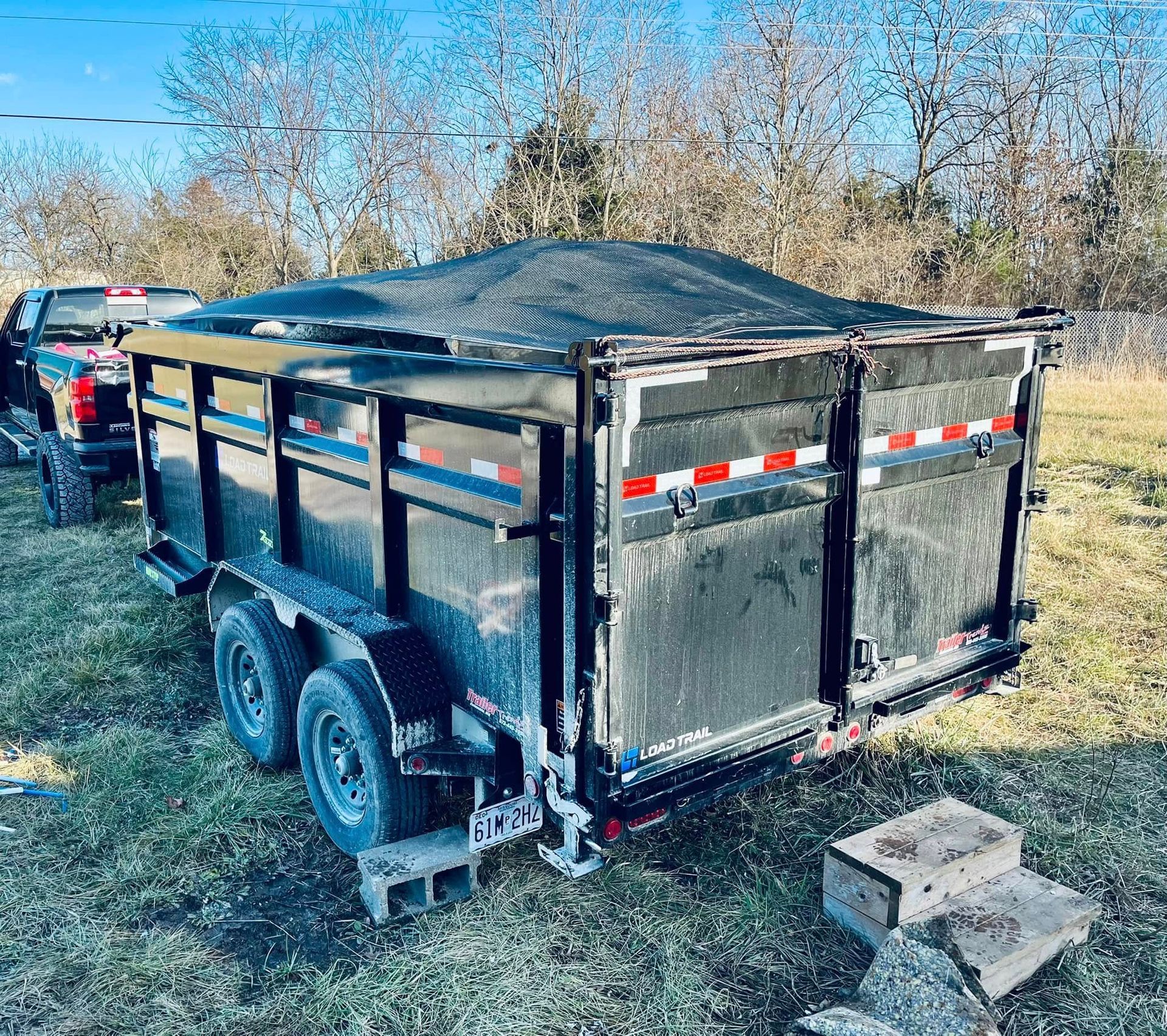 Black trailer with a mesh cover parked outdoors near a truck and some concrete blocks.