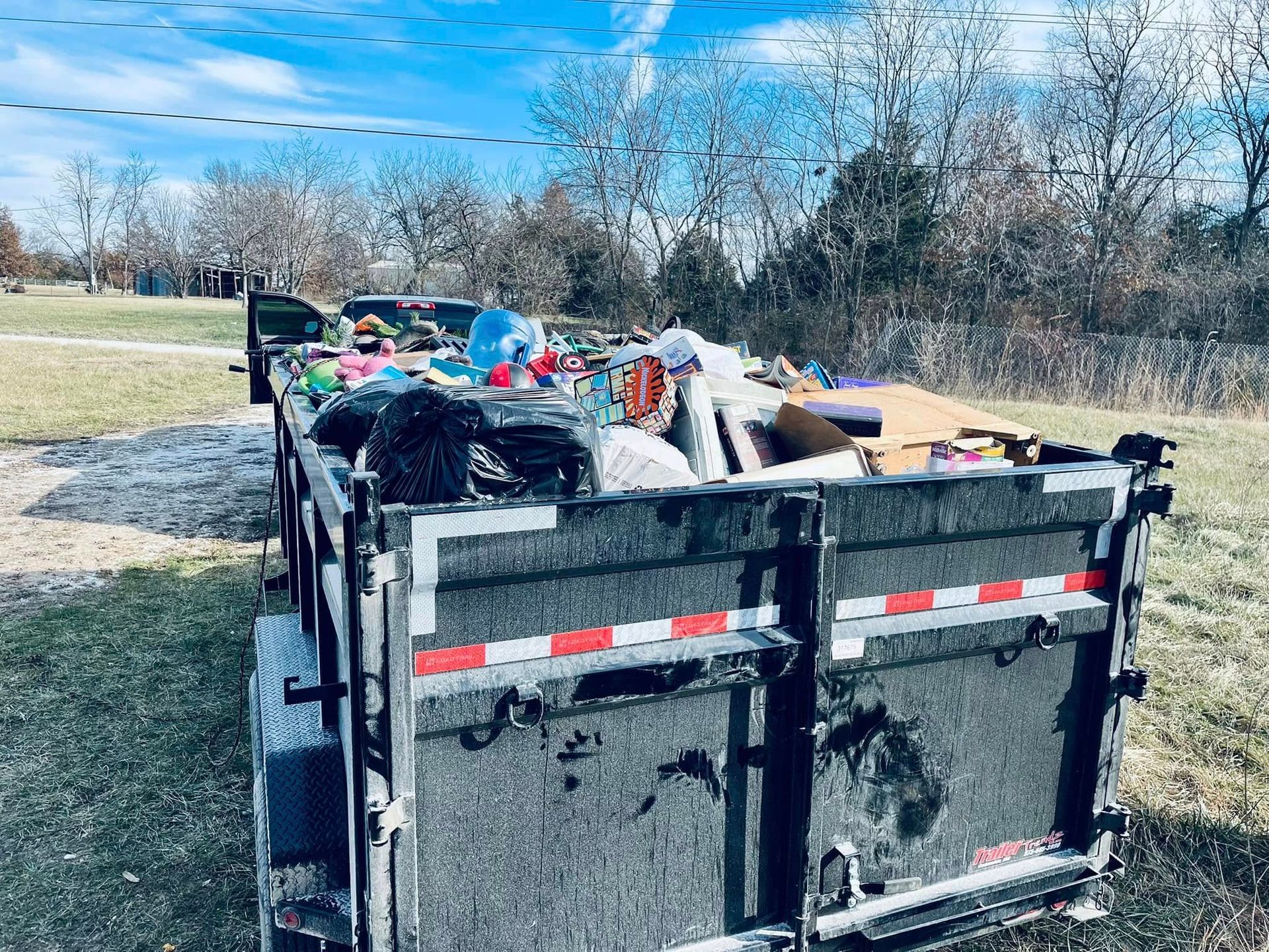 Black trailer overflowing with trash, in a grassy field on a sunny day.