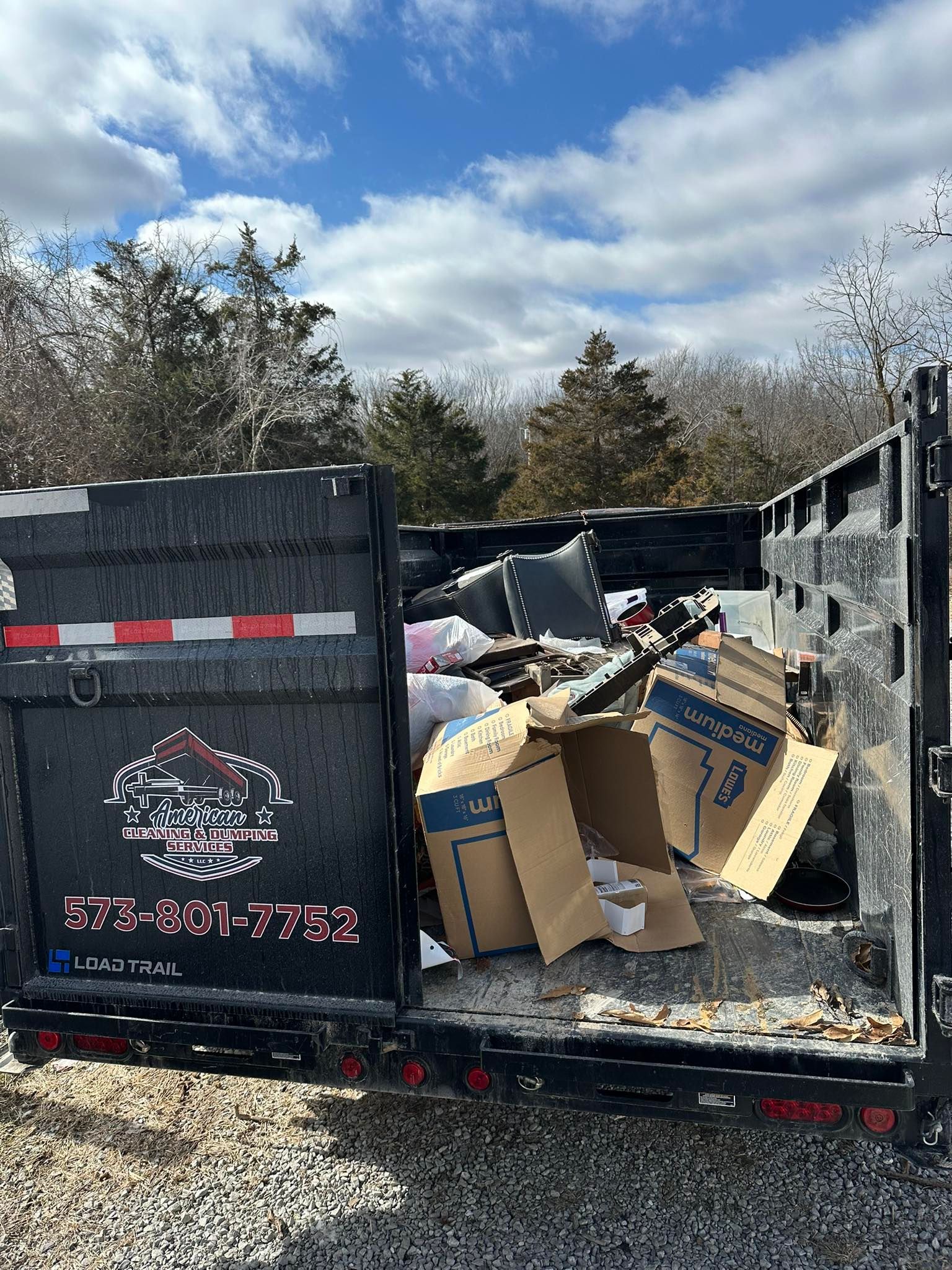 A black dumpster filled with trash and cardboard boxes against a cloudy sky.