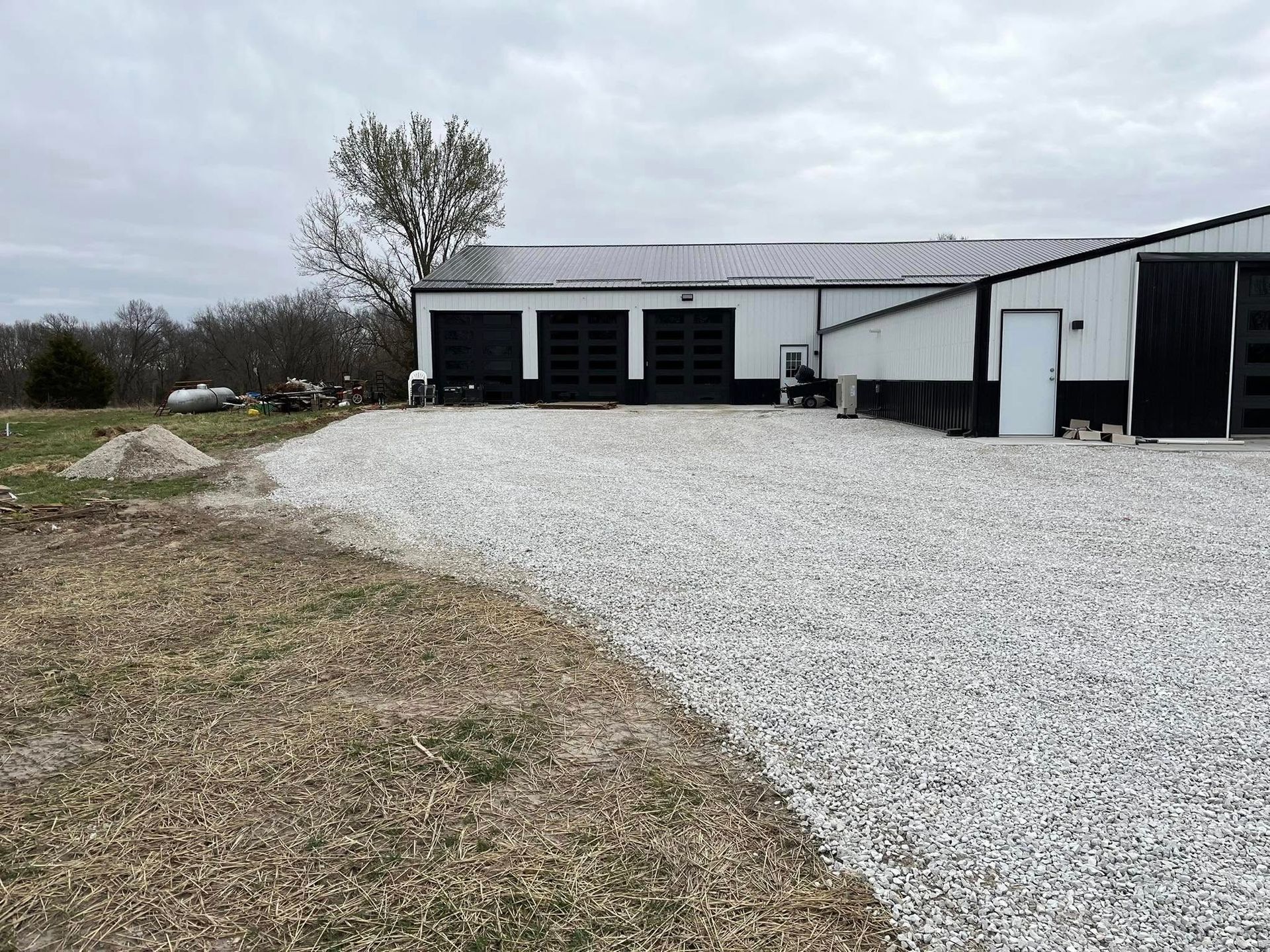 Gravel driveway in front of a white metal building with black garage doors. Overcast sky.