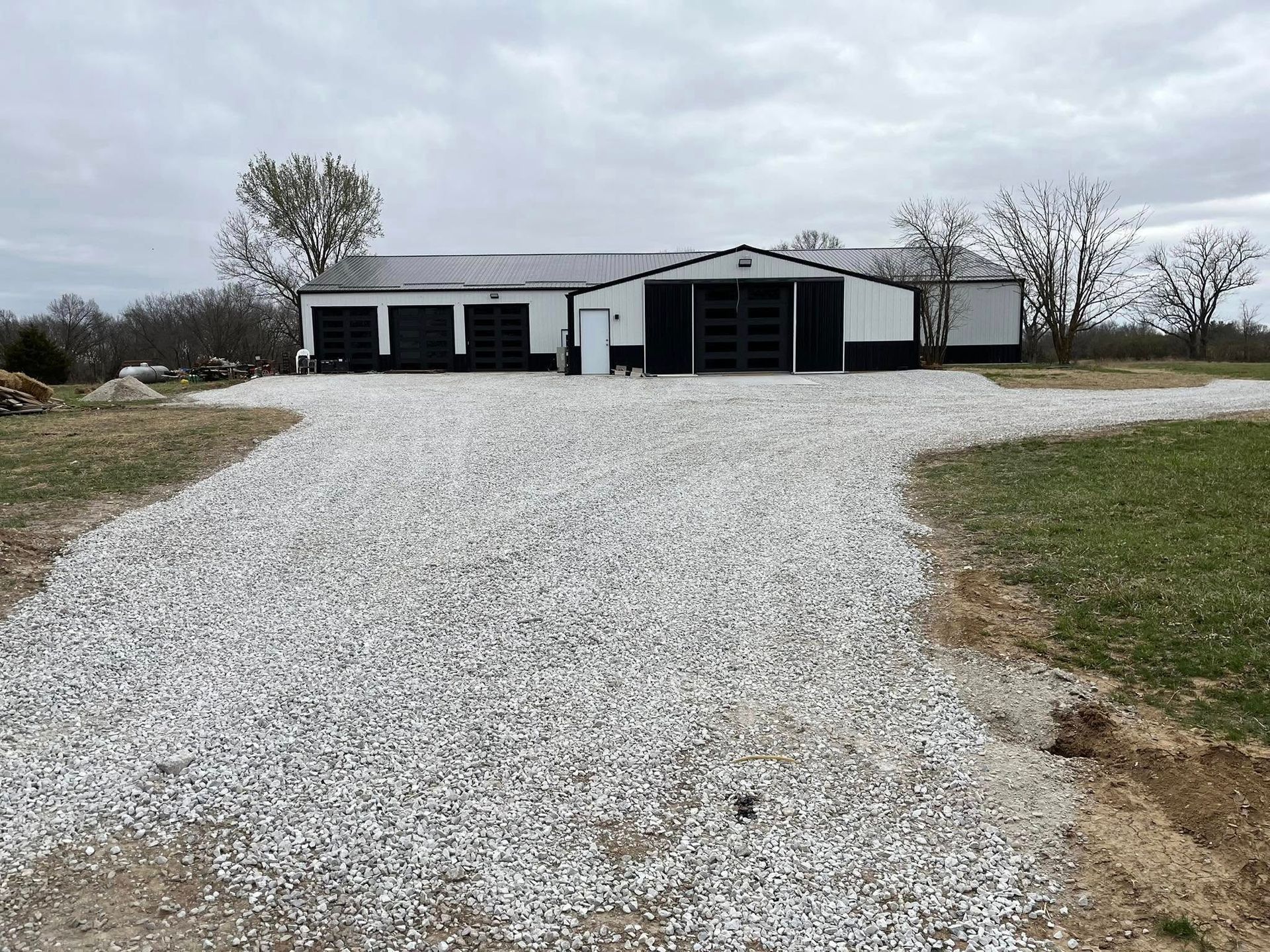 Gravel driveway leads to a black and white building with three garage doors and a door under an overcast sky.