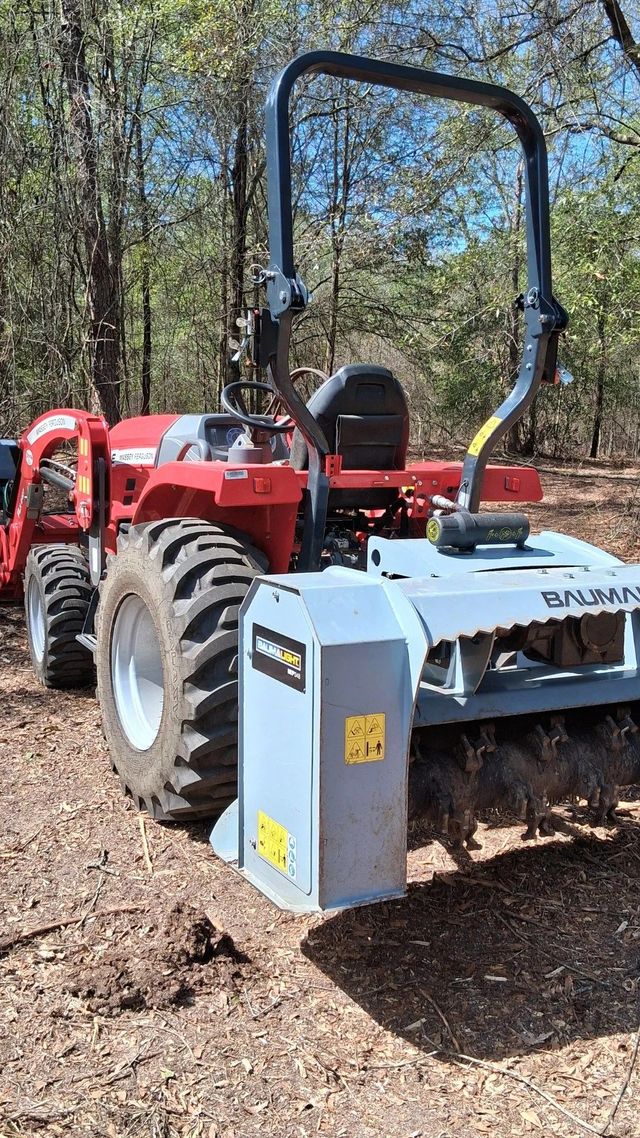 Red tractor with front loader and brush hog attachment parked on a wooded dirt path.