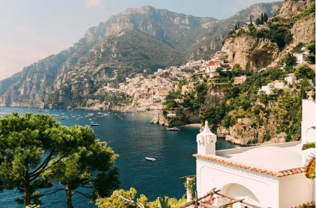 Coastal Italian town nestled into cliffs, with blue water and mountains in the background. Sunny day.