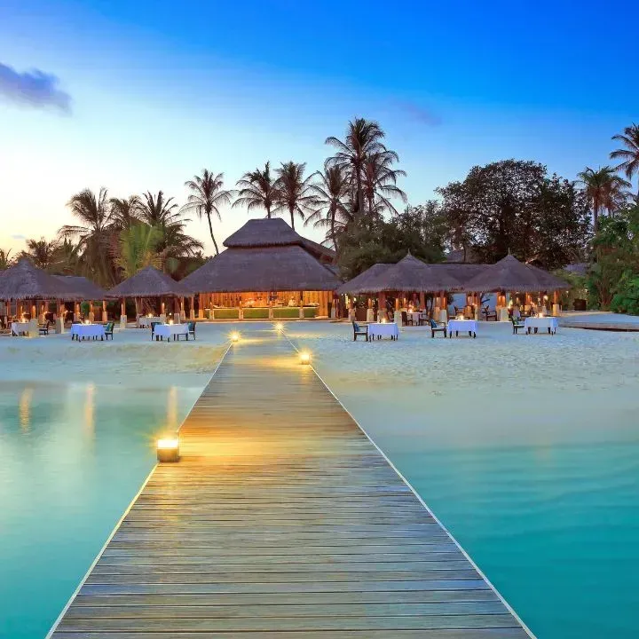Wooden pier leads to a beachside restaurant with tables set for dining at sunset with palm trees.