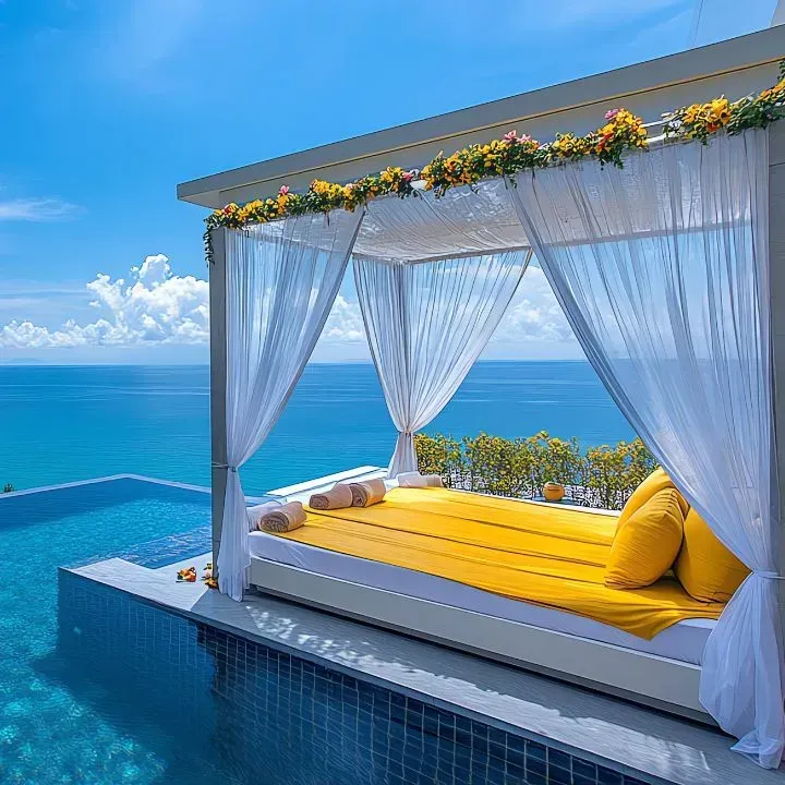 Outdoor daybed with yellow cushions and sheer white curtains, overlooking a pool and ocean.