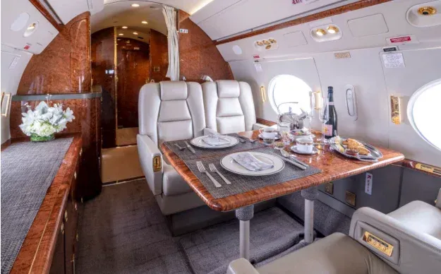 Interior of a private jet with a table set for dining, surrounded by cream-colored leather seats and wood paneling.