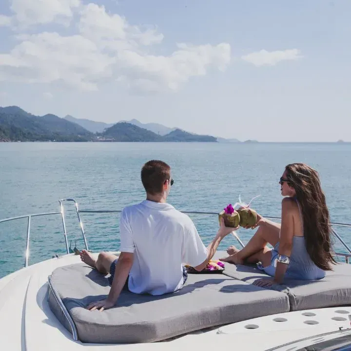 Couple relaxing on yacht, holding coconut drinks, ocean and mountains visible.