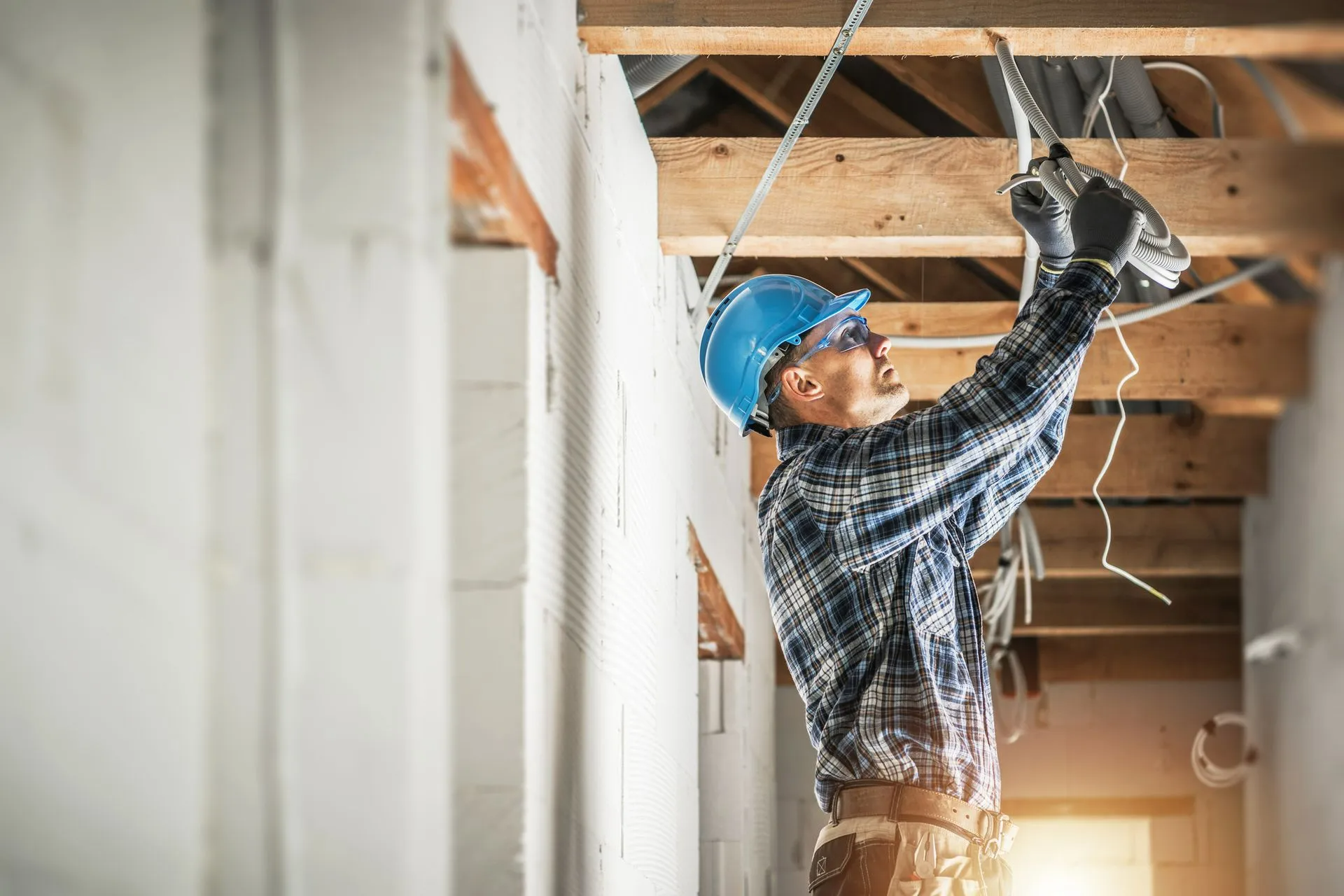 A worker in a hard hat and plaid shirt installs electrical wiring in the exposed ceiling of a construction site.