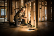 A person in a hard hat kneeling on a concrete floor to install electrical wiring in the wood frame of a building.