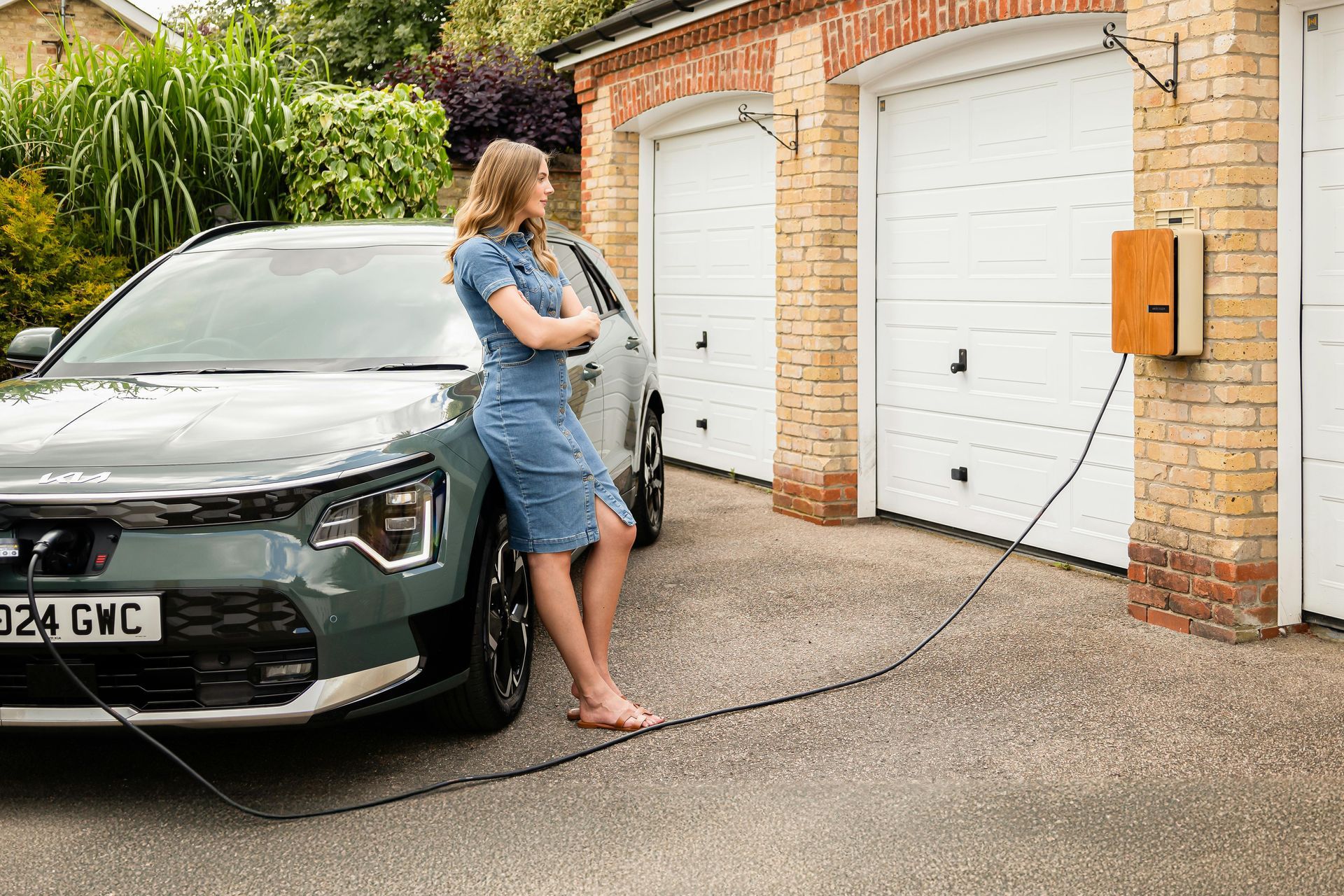 A person leans against a grey electric vehicle parked in a gravel driveway, charging via a wall-mounted unit.