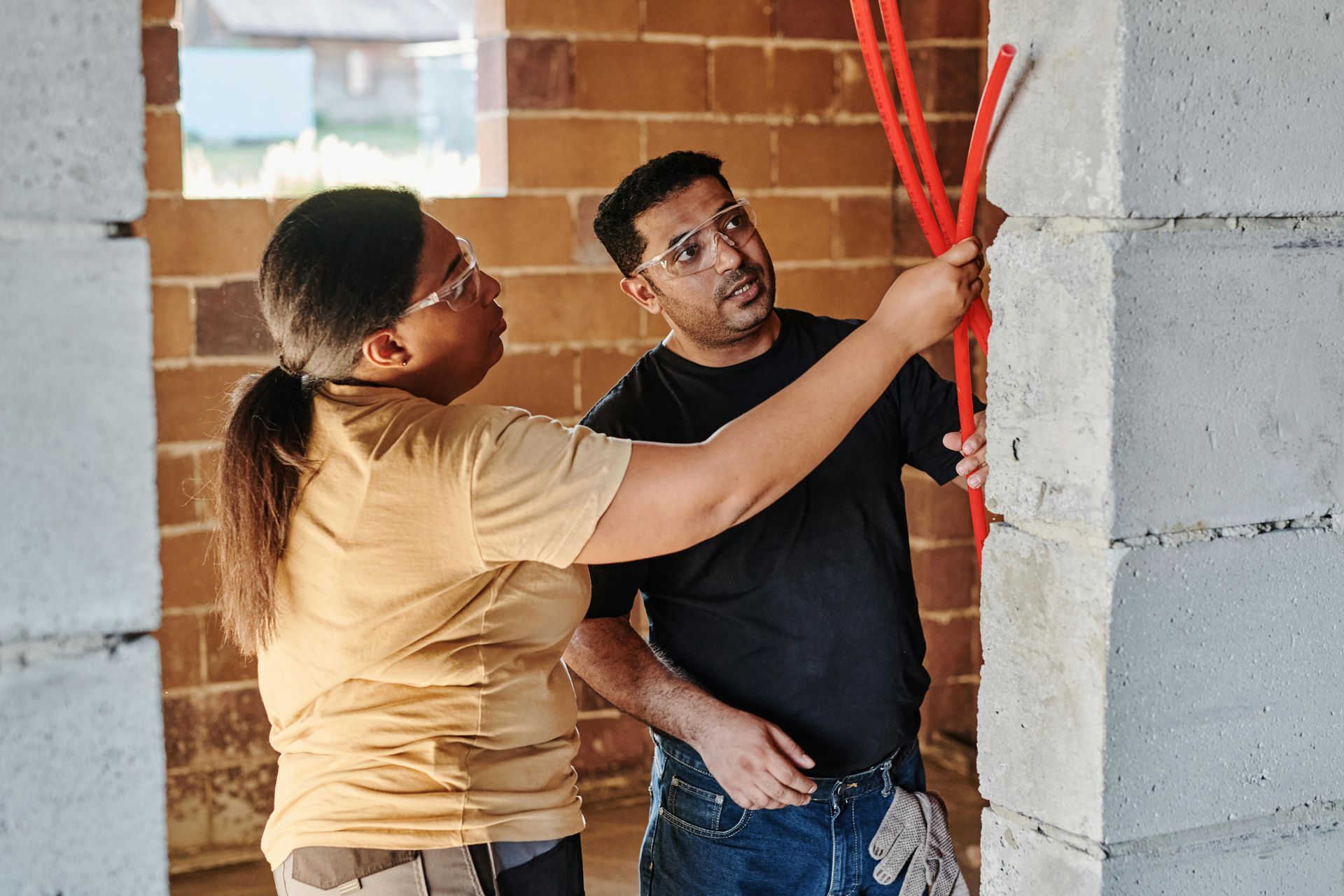 Two people in a construction site examine red cables extending from a concrete block wall.