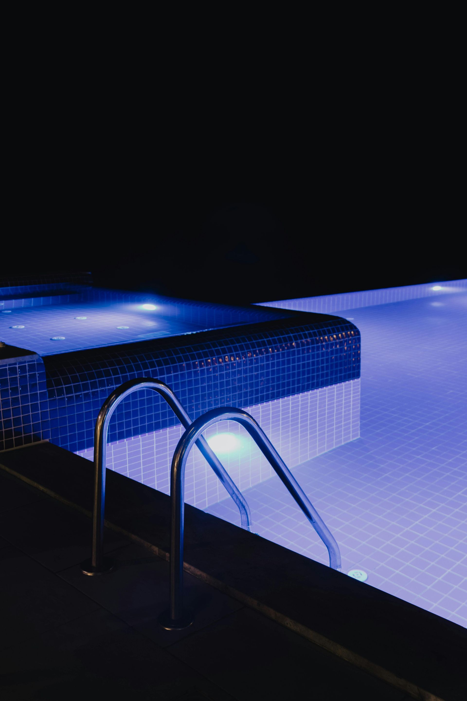 A swimming pool and attached hot tub illuminated with blue underwater lights at night, featuring a stainless steel ladder.