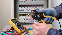 An electrician in gloves uses wire strippers near a circuit breaker panel with a multimeter on the table.