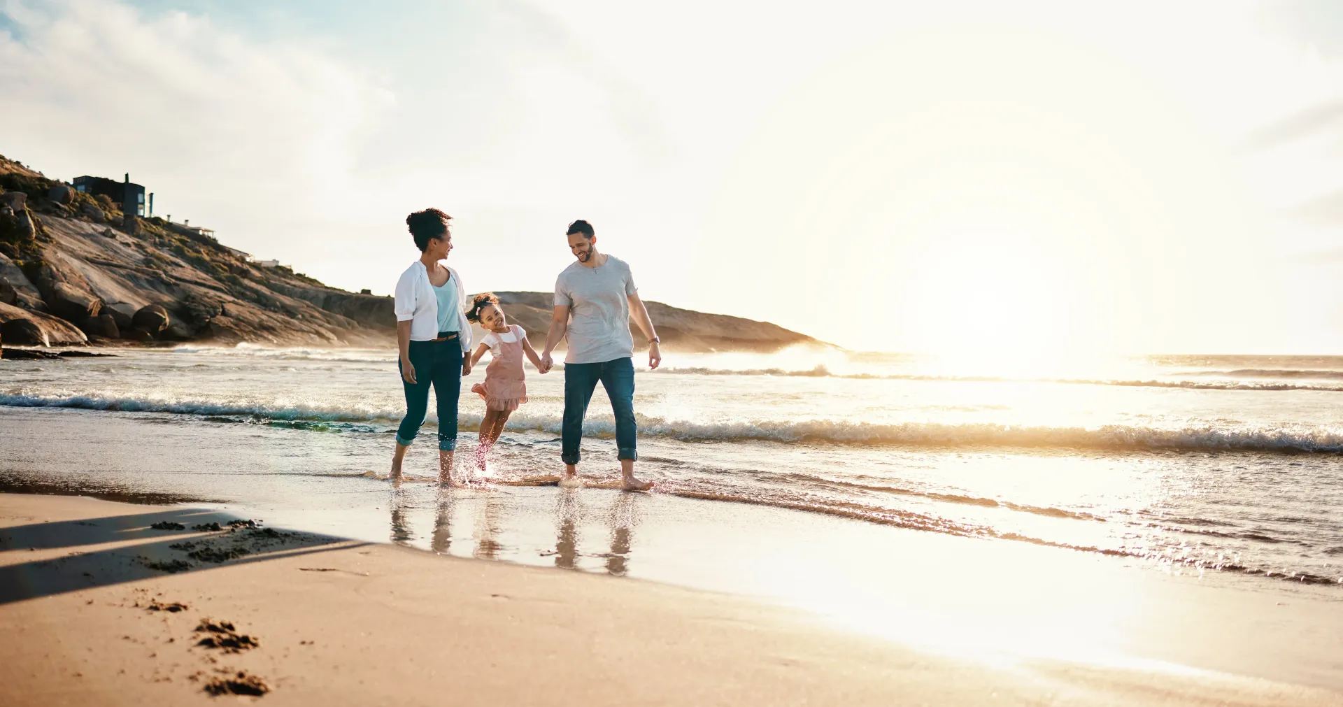 A family walks along the shoreline at sunset, holding hands as they play in the shallow surf.