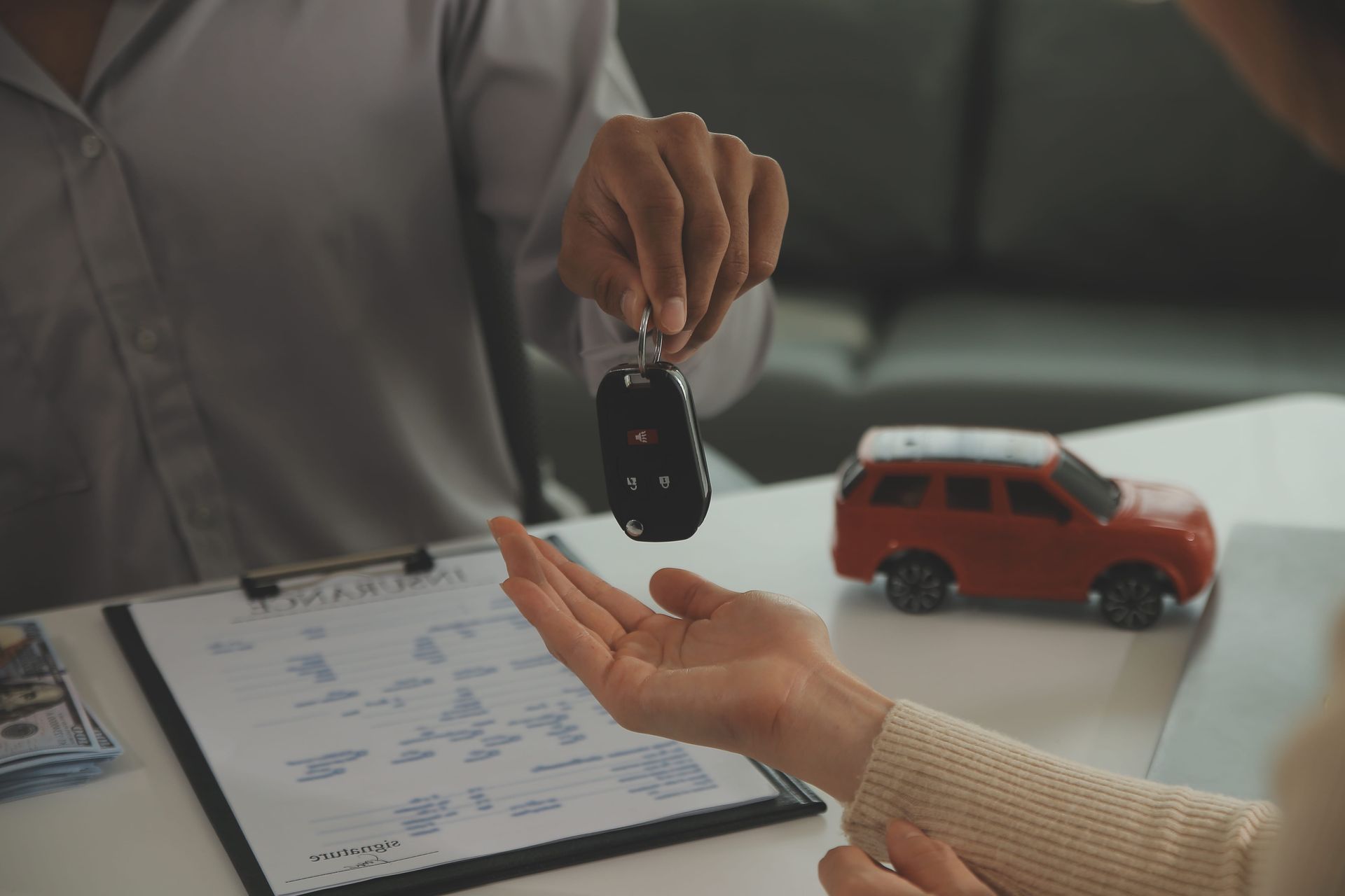 Car salesperson handing keys to a customer beside a red toy car and paperwork on a desk