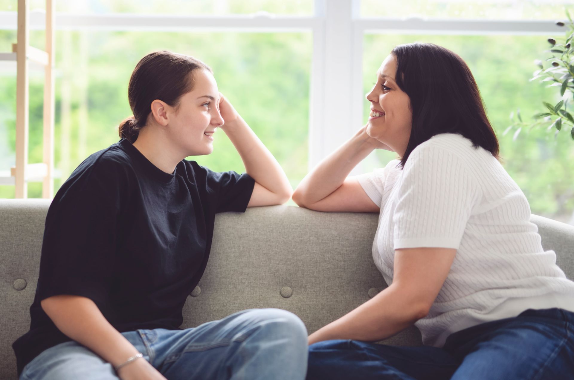 Two people sitting on a couch, smiling and talking by a bright window