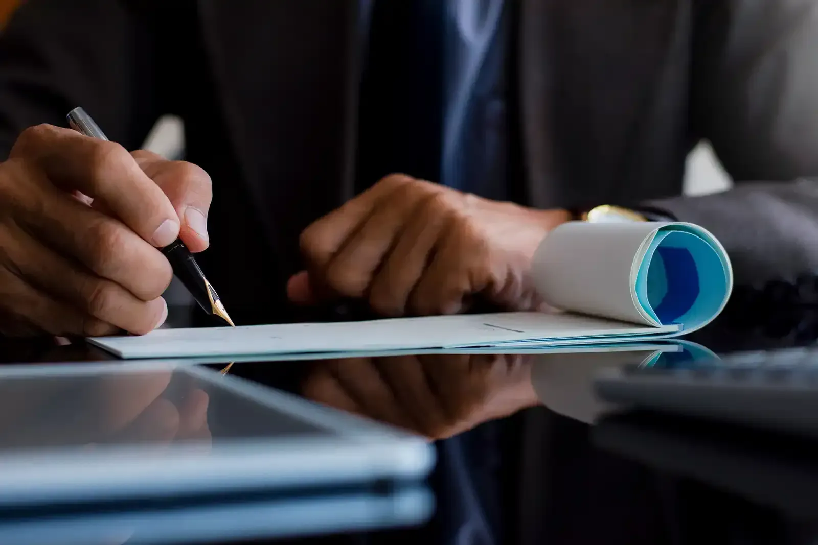 Hands writing on paper beside a rolled blueprint on a table