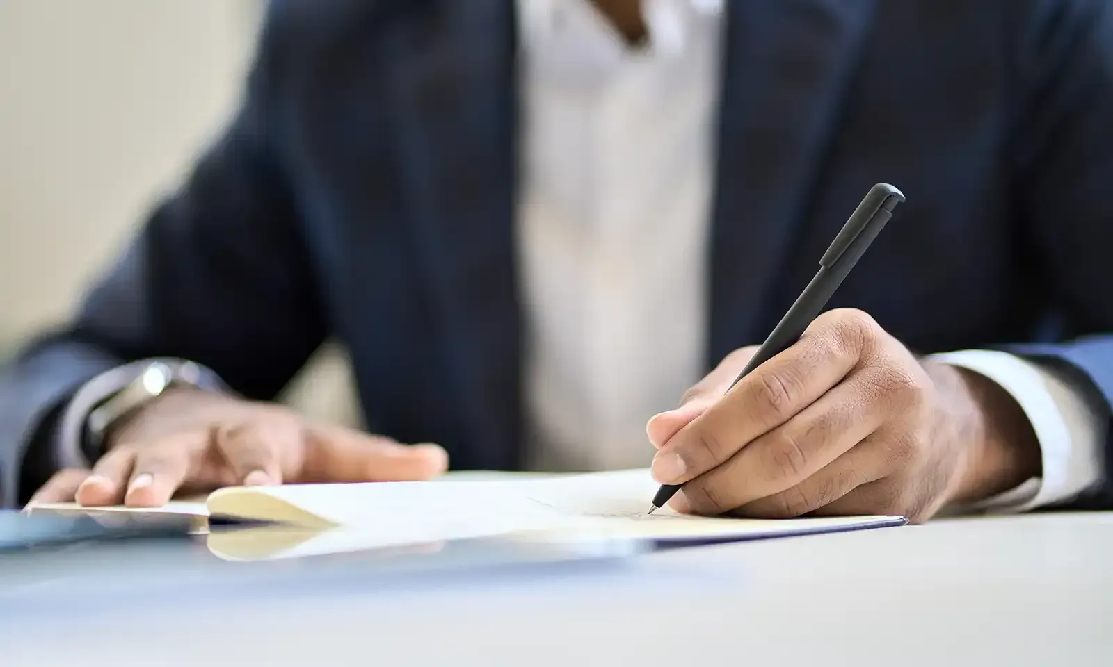 A person in a dark suit writing in a notebook with a black pen at a desk.