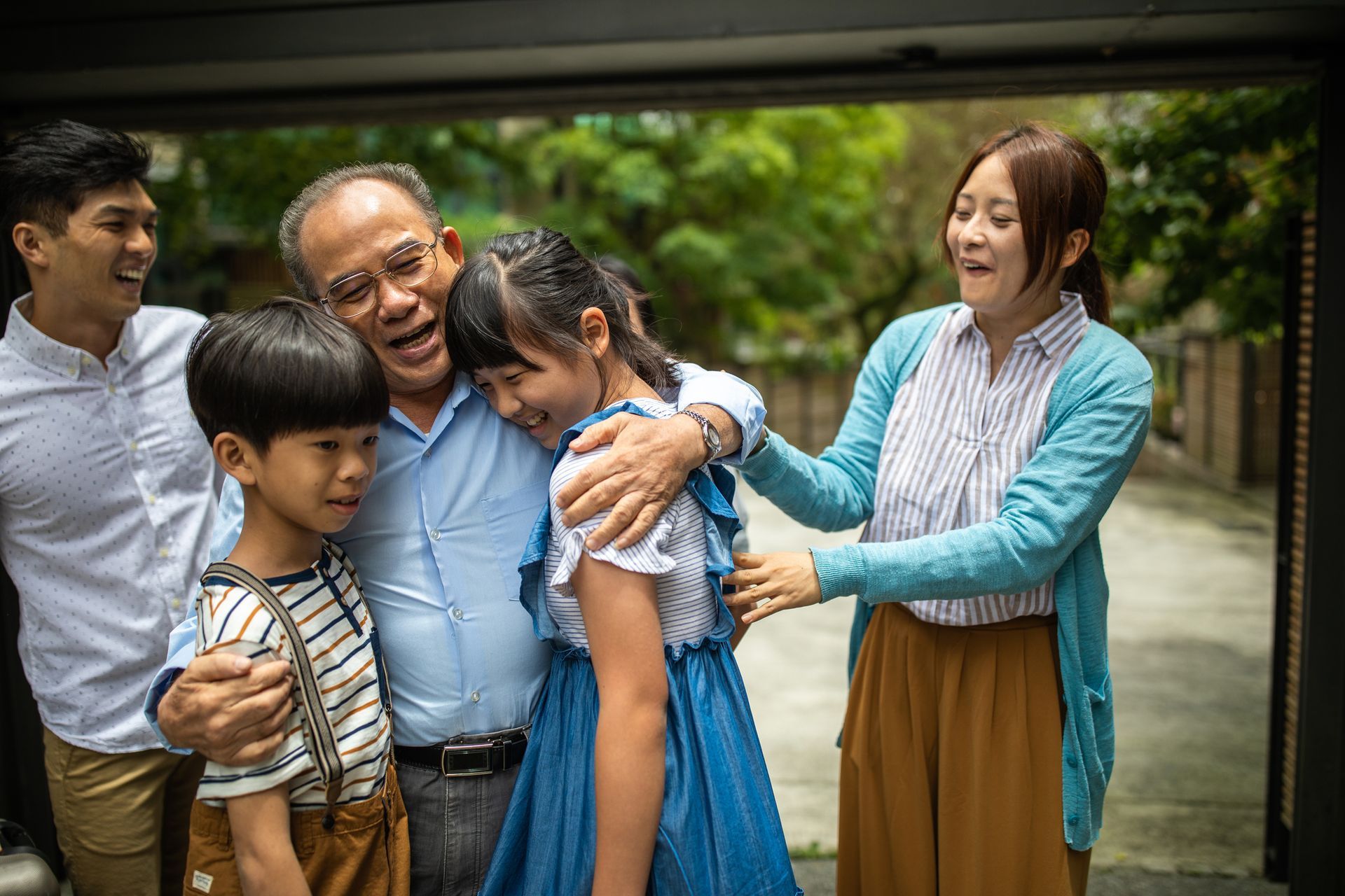 A multi-generational family smiles and embraces while standing together outdoors.