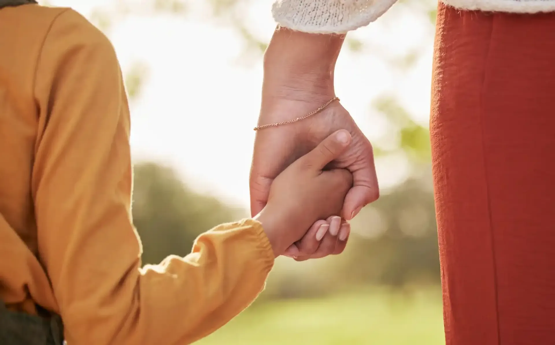Close-up of a person holding a smaller hand in a gentle, supportive grip, against a soft, blurred outdoor background.