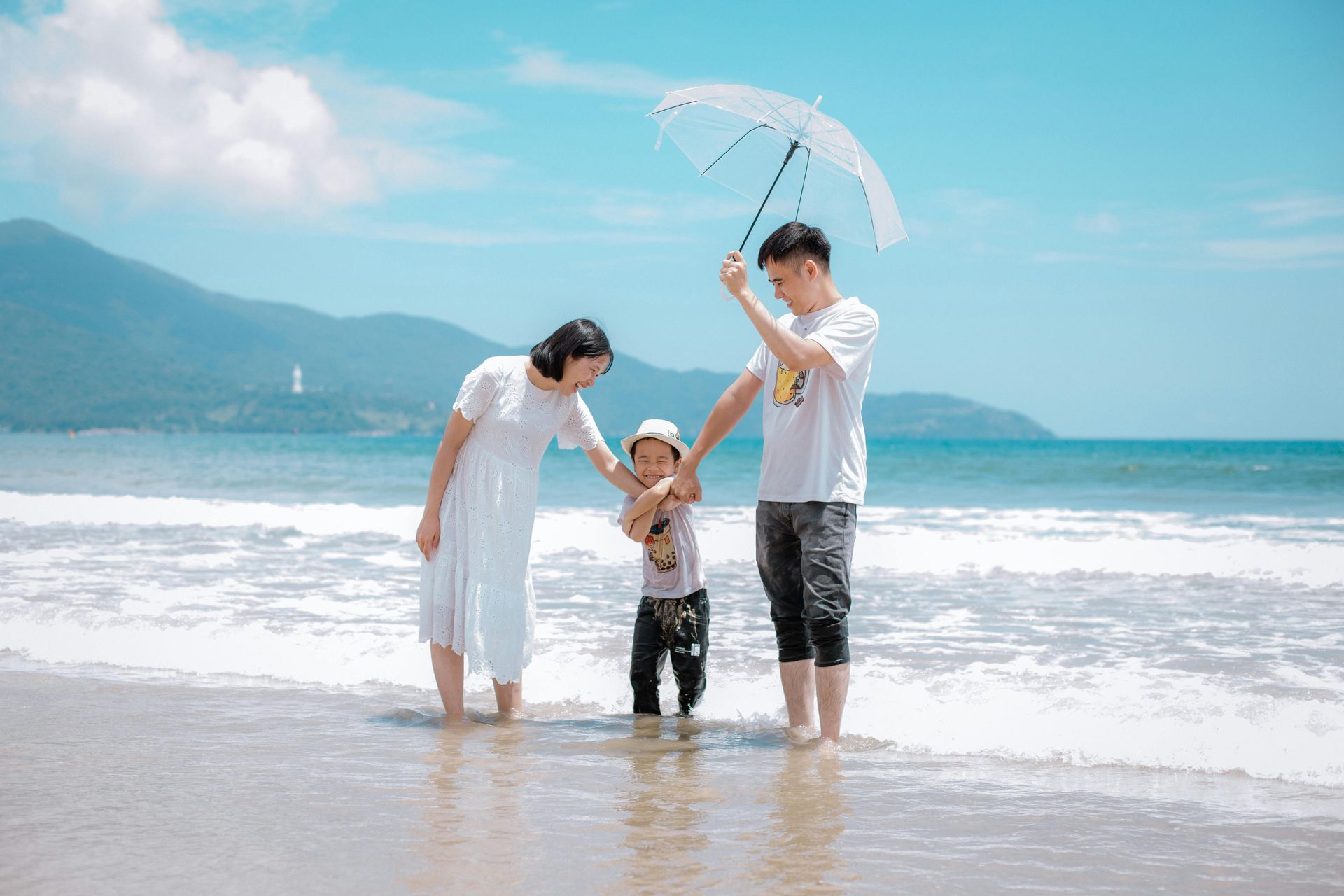 A family of three stands in shallow ocean waves on a sunny beach, holding hands under a single white umbrella.