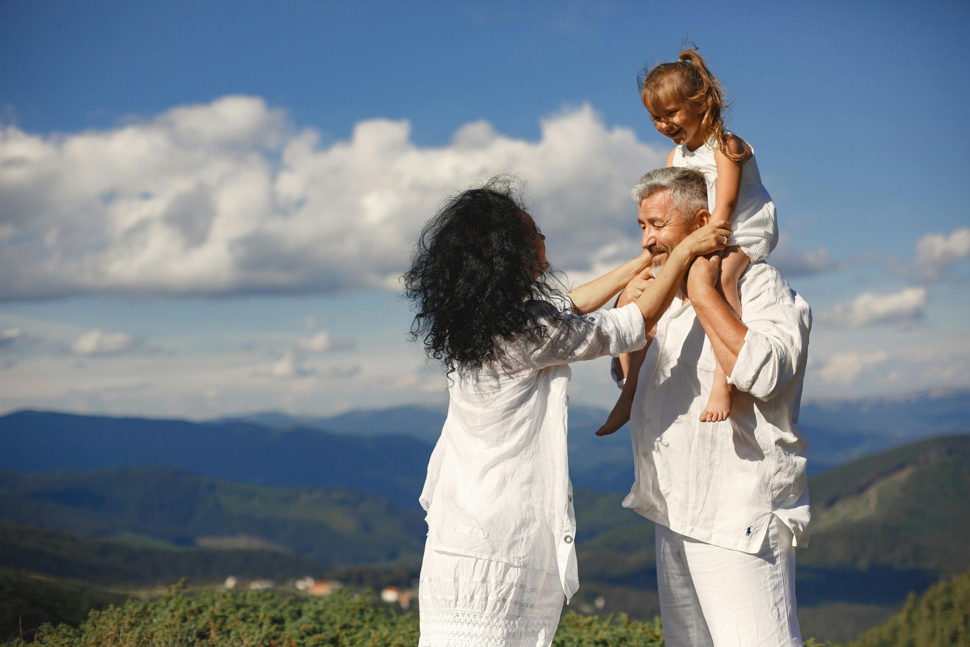 A family in white clothes, with a child on shoulders, smiles while standing outdoors against a mountain landscape.