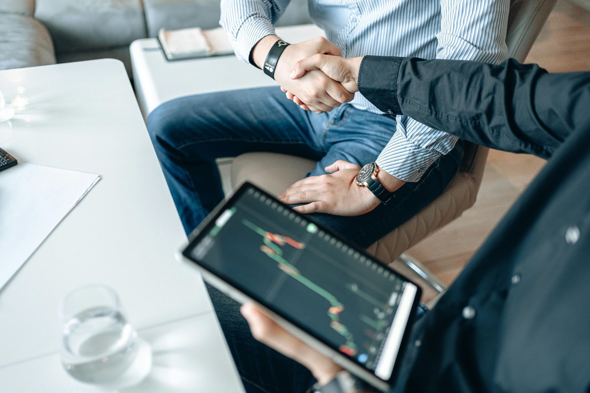 Two people in business attire shake hands over a tablet displaying financial stock market charts in an office.