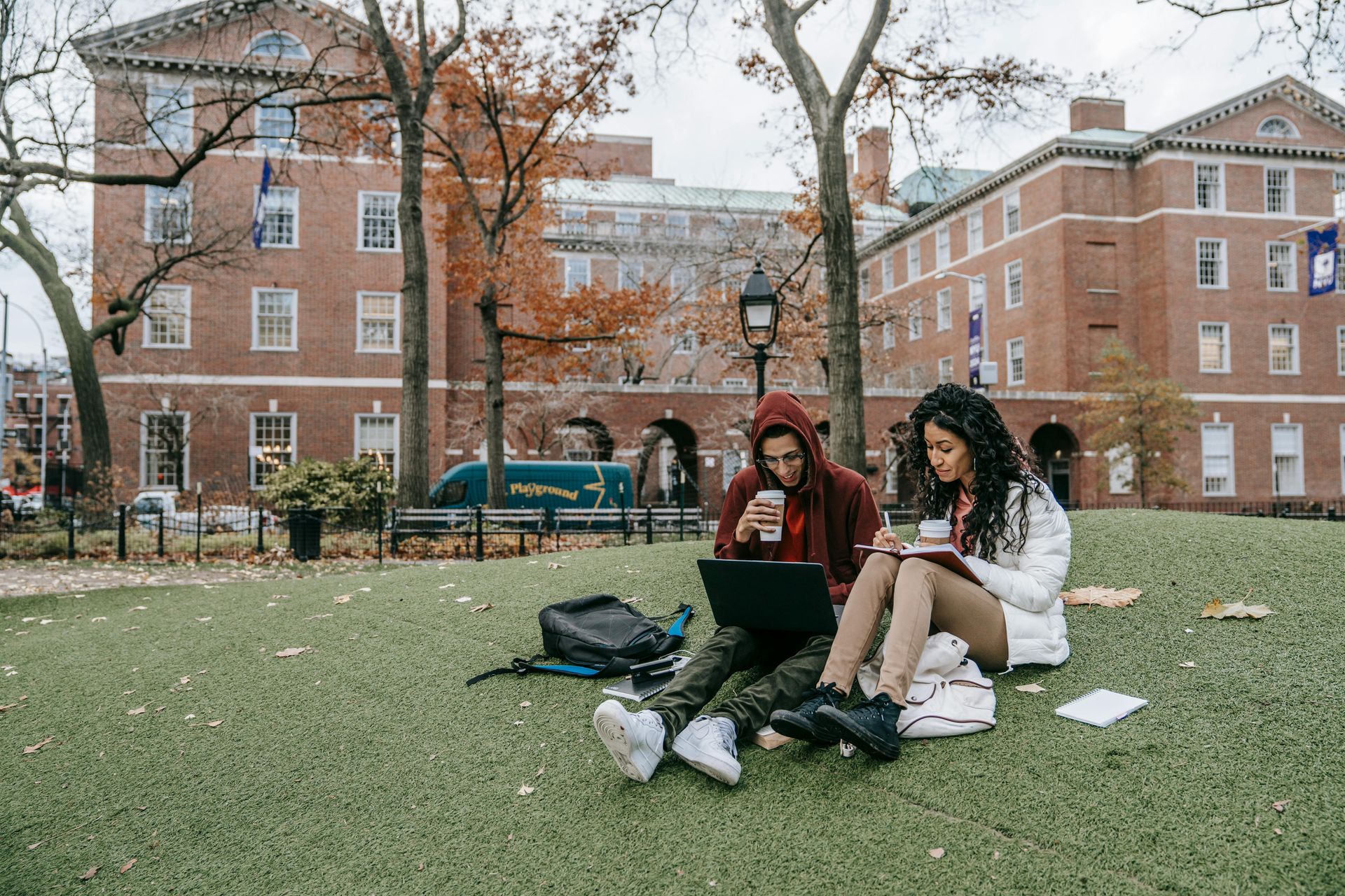 Two students sit on a grassy campus lawn, one using a laptop and the other writing in a notebook near brick buildings.