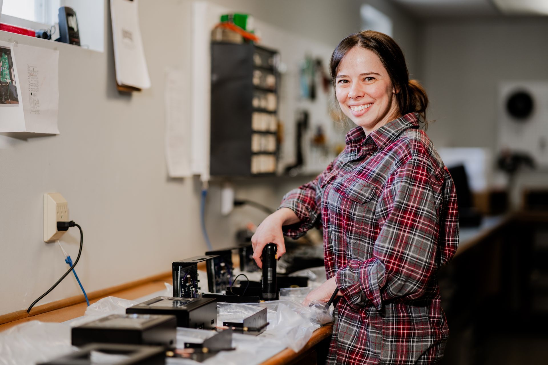 Woman in plaid shirt smiles while working on machinery at a workbench. | Photos by Kodjoarts
