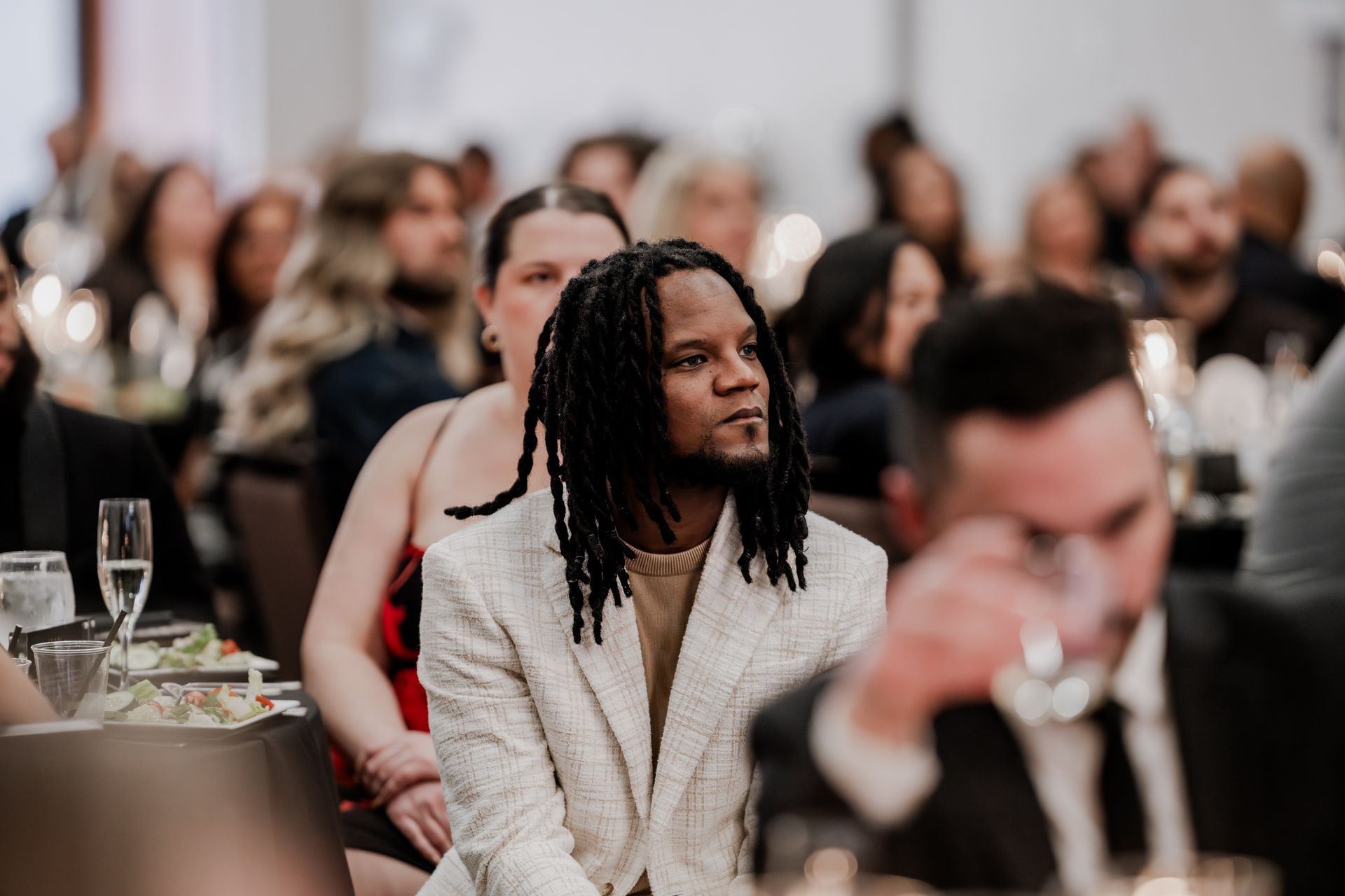 A person with locs in a cream blazer sits at a table during a crowded event, looking toward the side with focus.