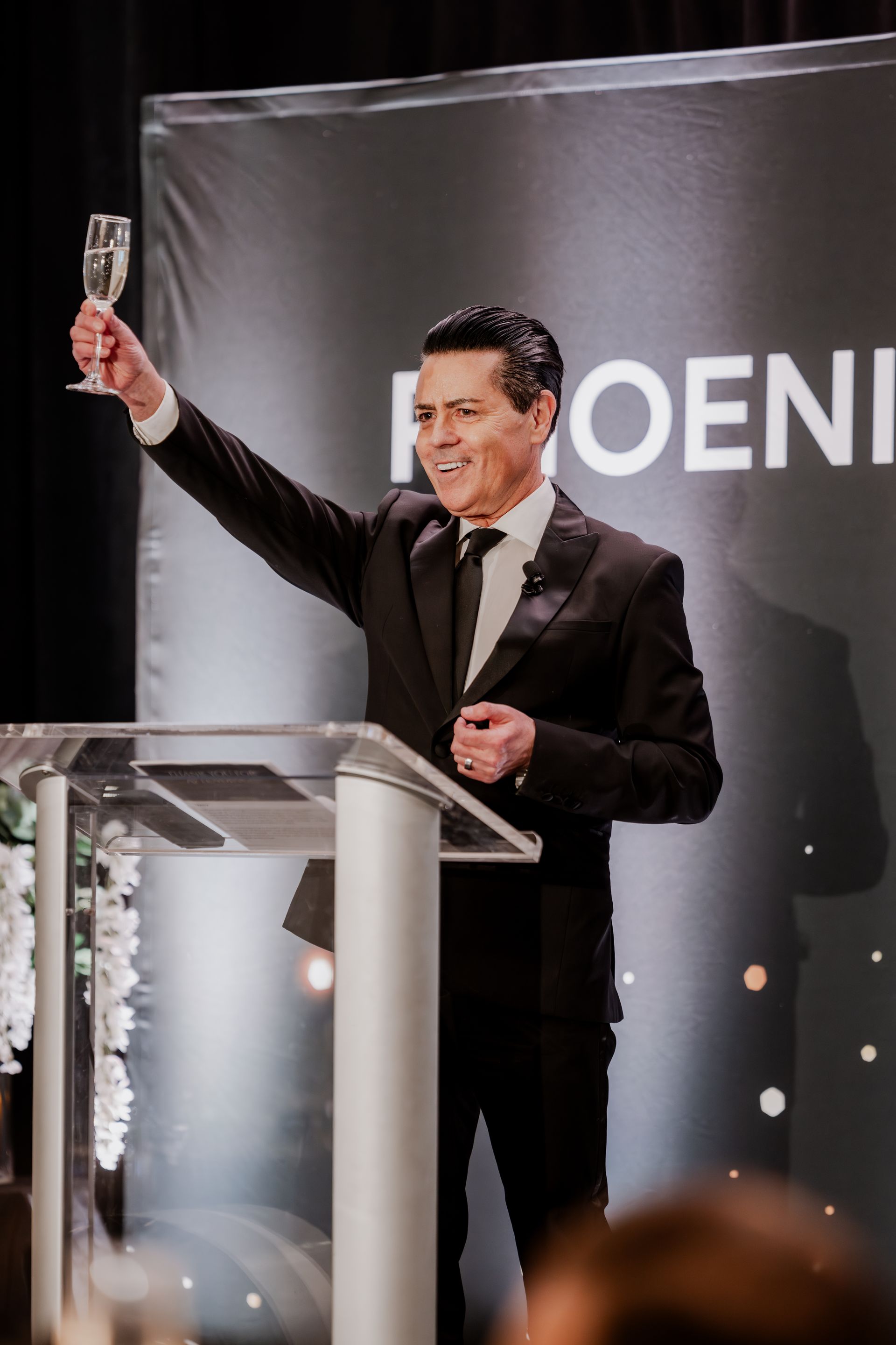 A person in a black suit stands at a podium, smiling and holding a champagne flute aloft for a toast in front of a banner.