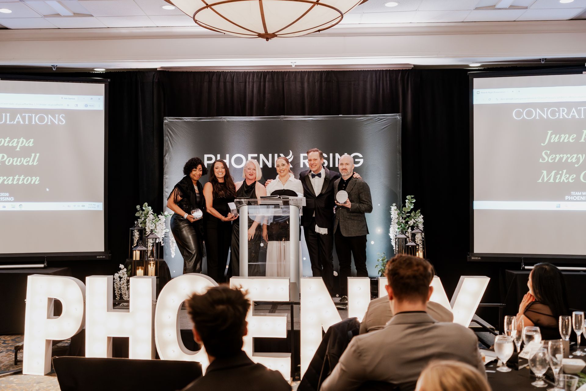 A group of people standing on a stage behind a podium under a Phoenix Rising sign, with large light-up letters in front.