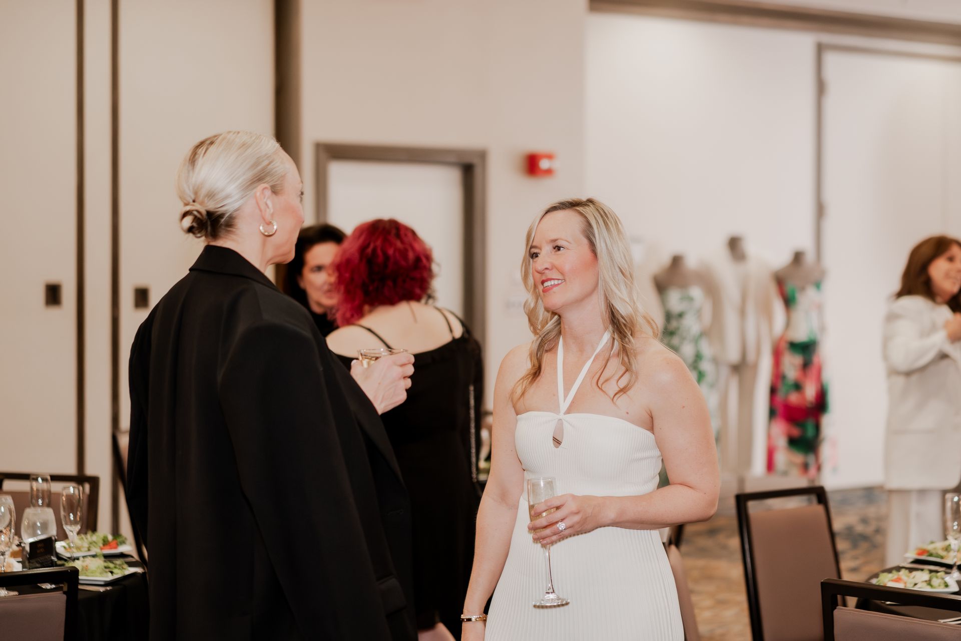 People socialize at a formal event, with two women in the foreground conversing while holding drinks.