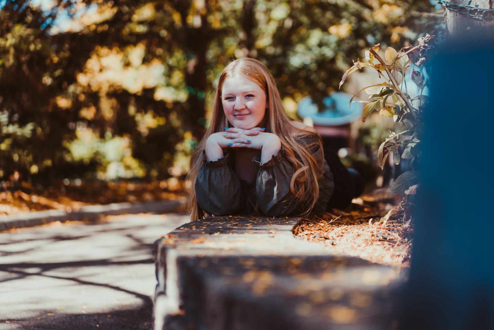 A high school senior is sitting on a bench in a park with her hands on her chin .