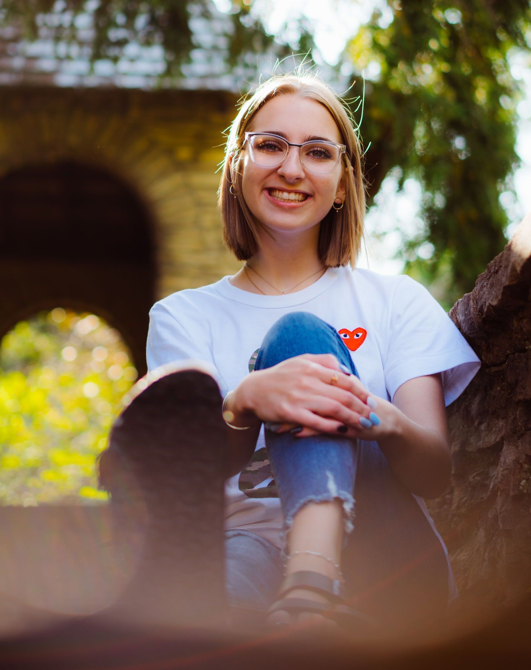 High school senior wearing glasses is sitting on a tree branch with her legs crossed .