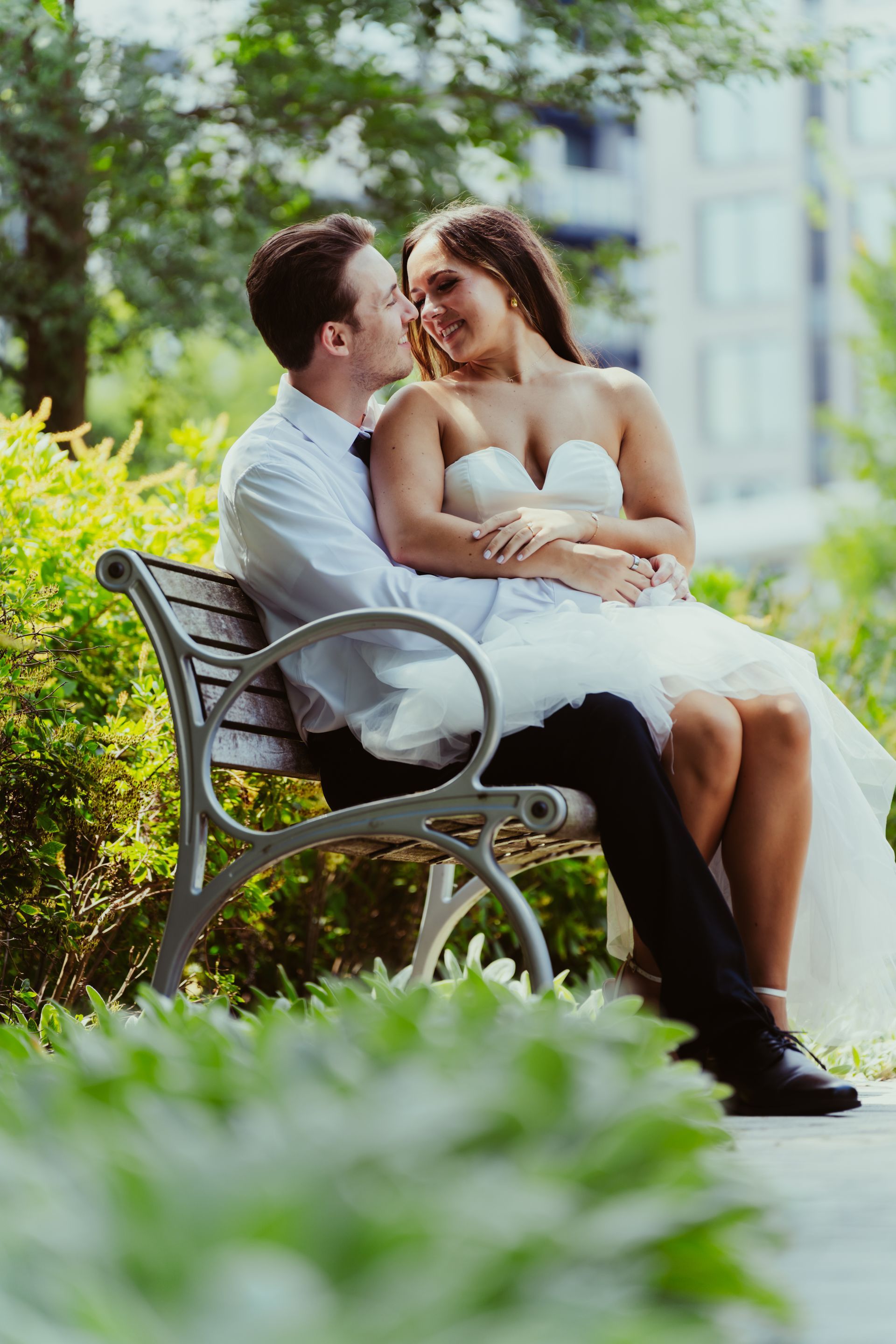 A bride and groom are sitting on a park bench and kissing.