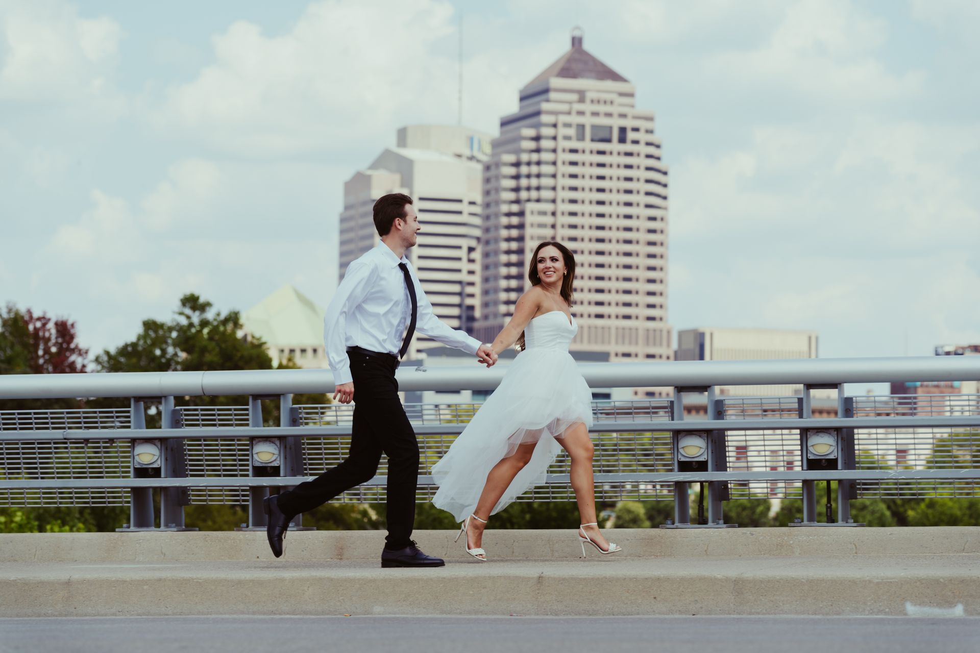 A bride and groom are running across a bridge holding hands