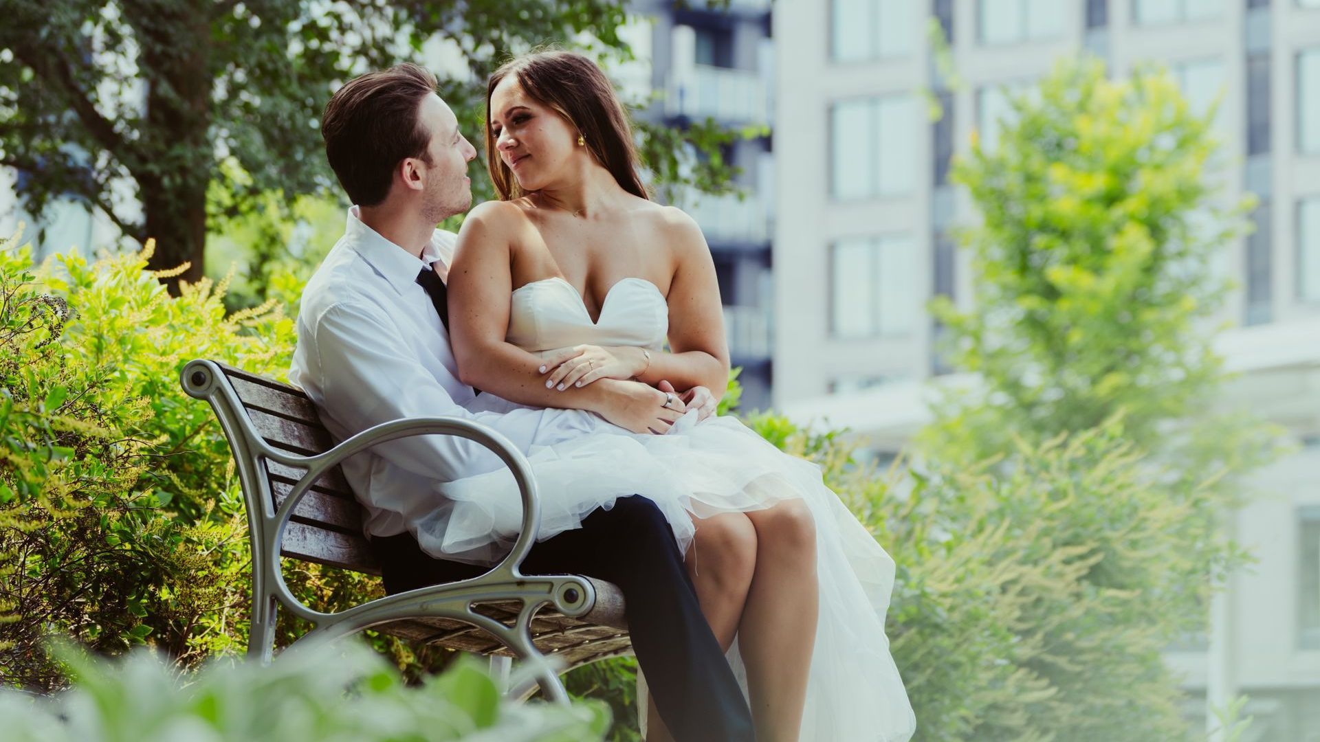 A bride and groom are kissing on a park bench.