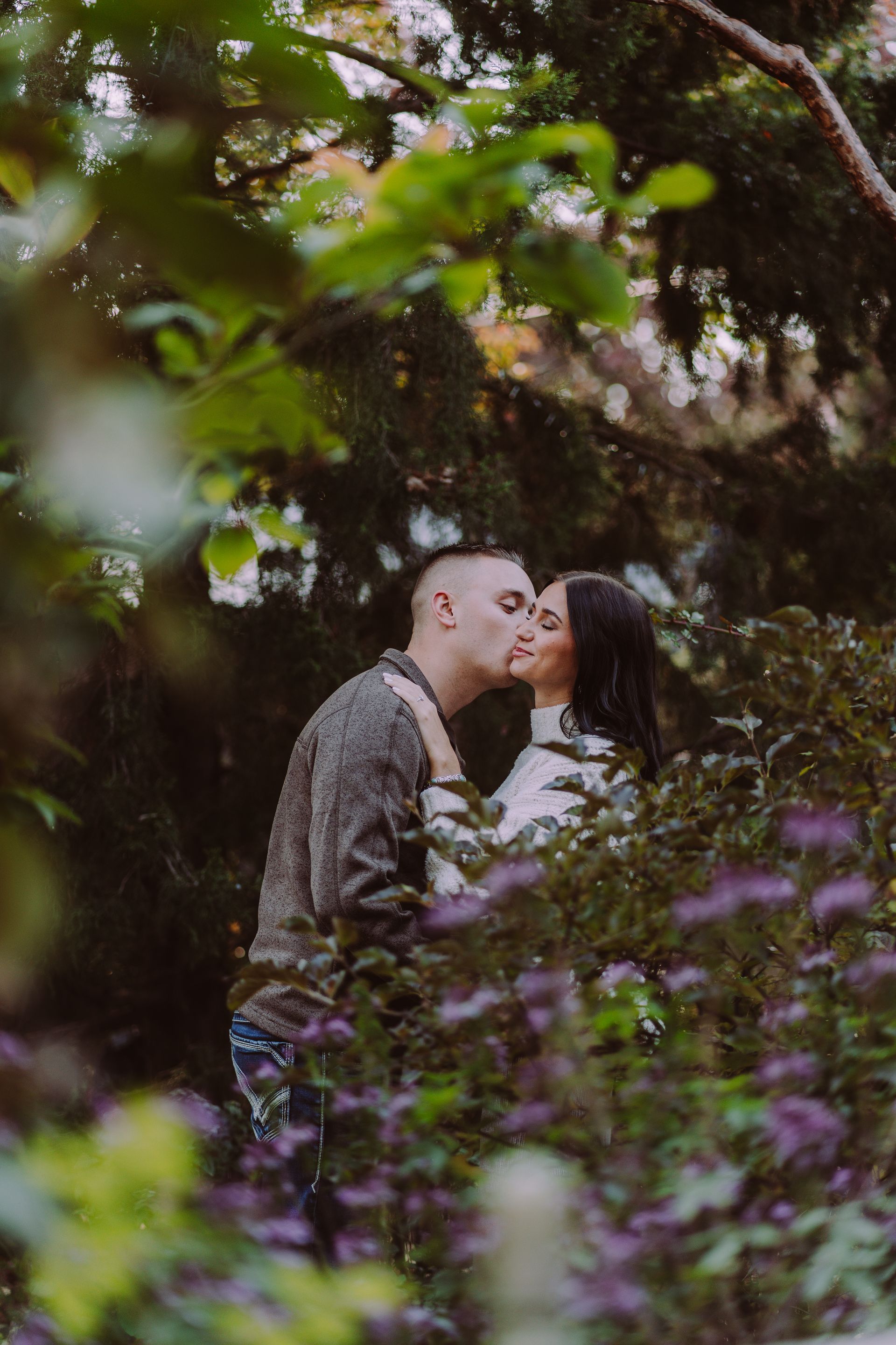 Couple kissing amidst lush foliage, natural lighting.