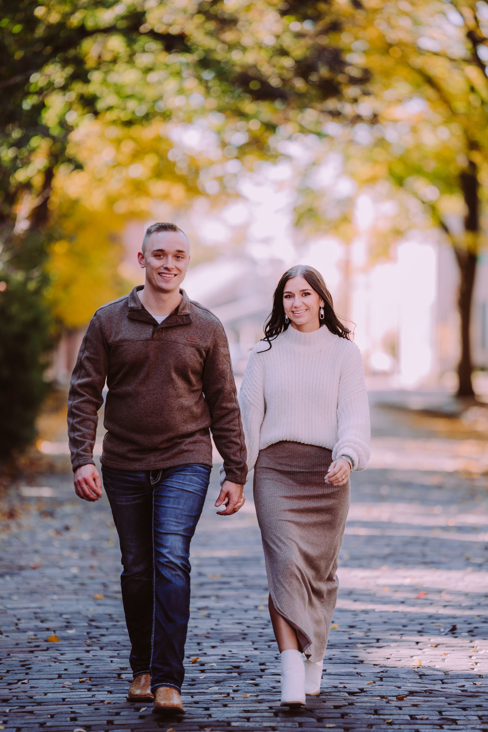 Couple walking hand-in-hand on cobblestone path under autumn trees. Man smiles, woman looks forward.