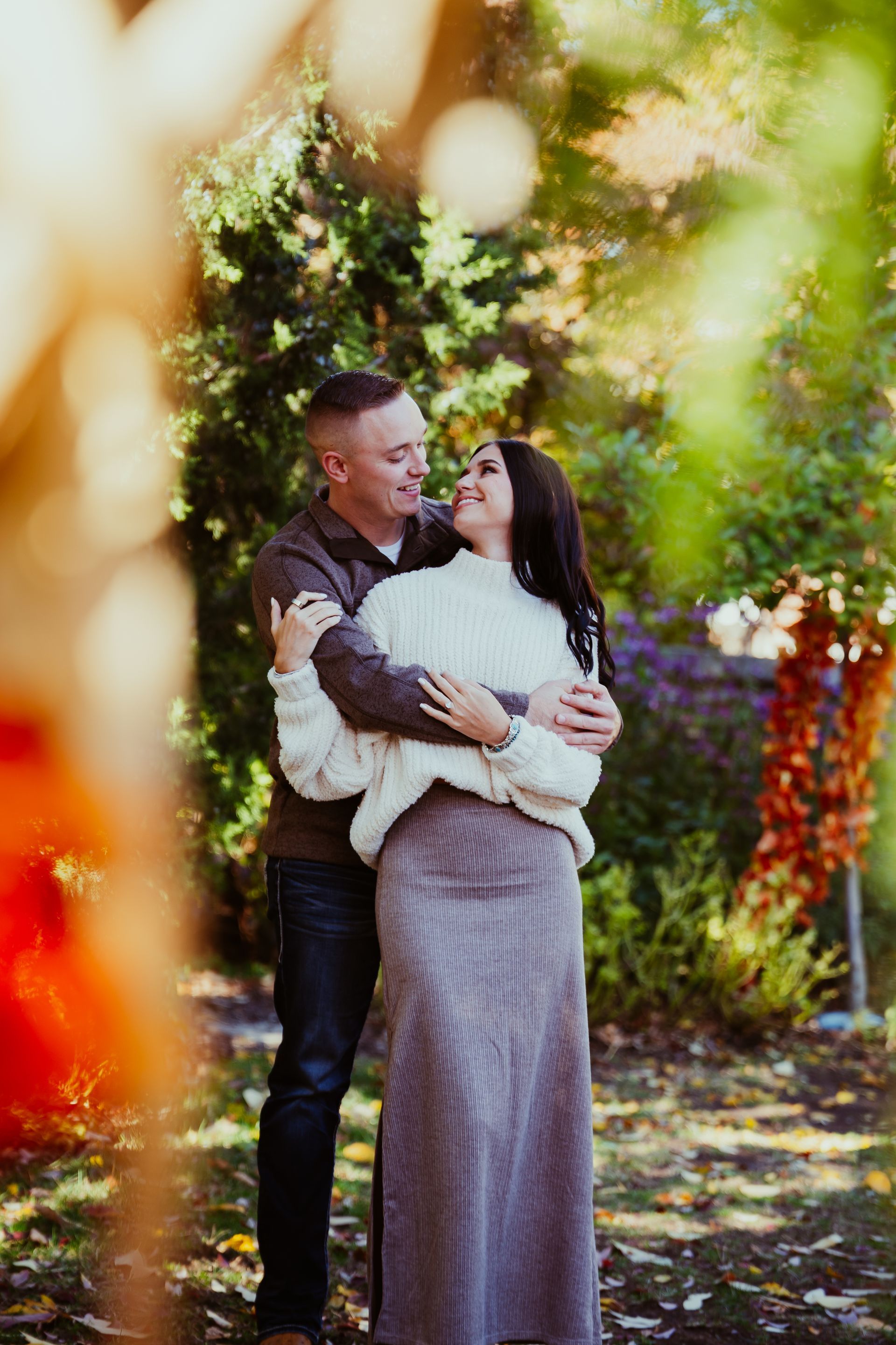 Couple embracing in a garden, smiling at each other. Man in brown, woman in cream sweater and gray skirt. Autumn leaves in foreground.