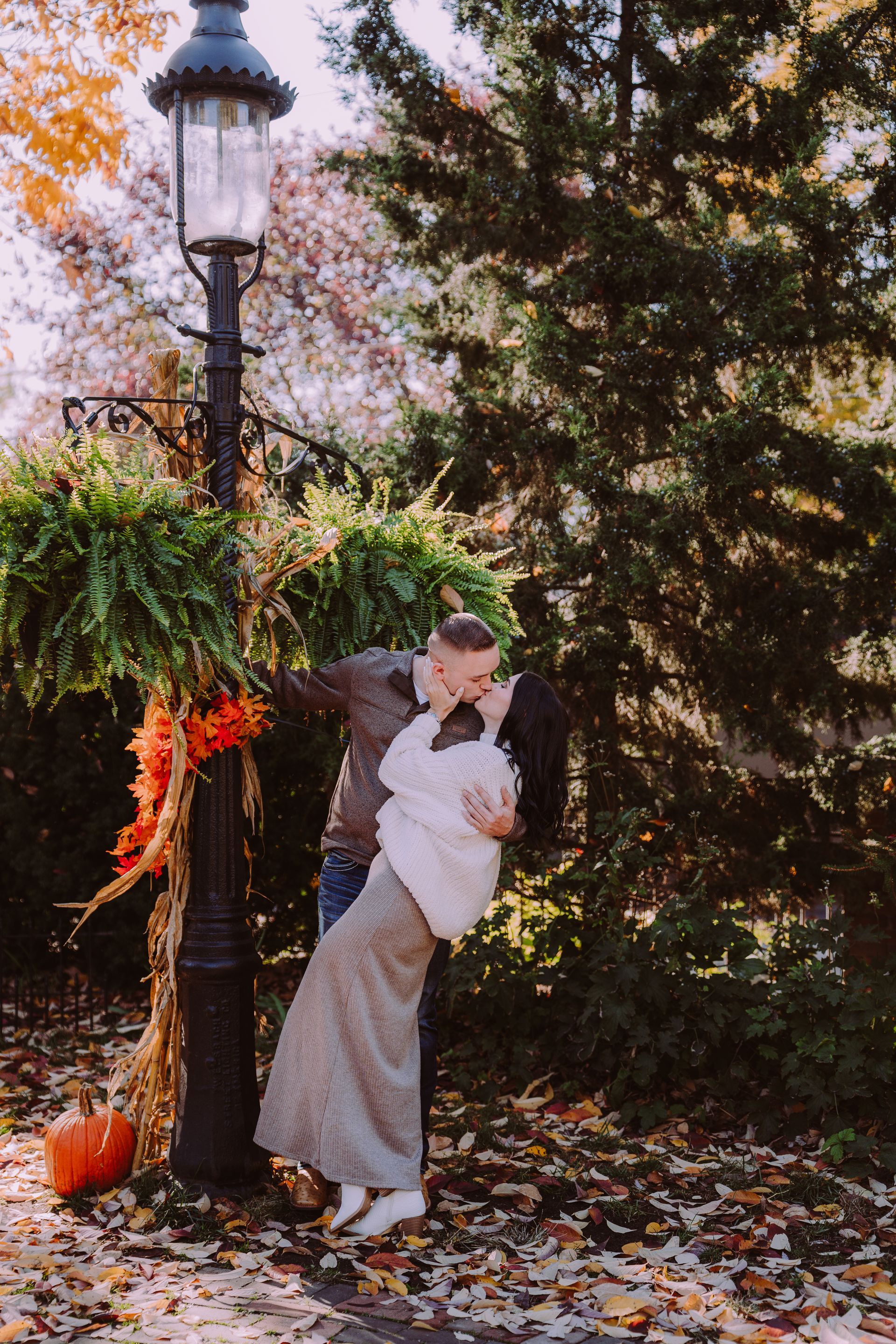 Couple kissing near a lamp post decorated with fall foliage. Leaves on the ground.