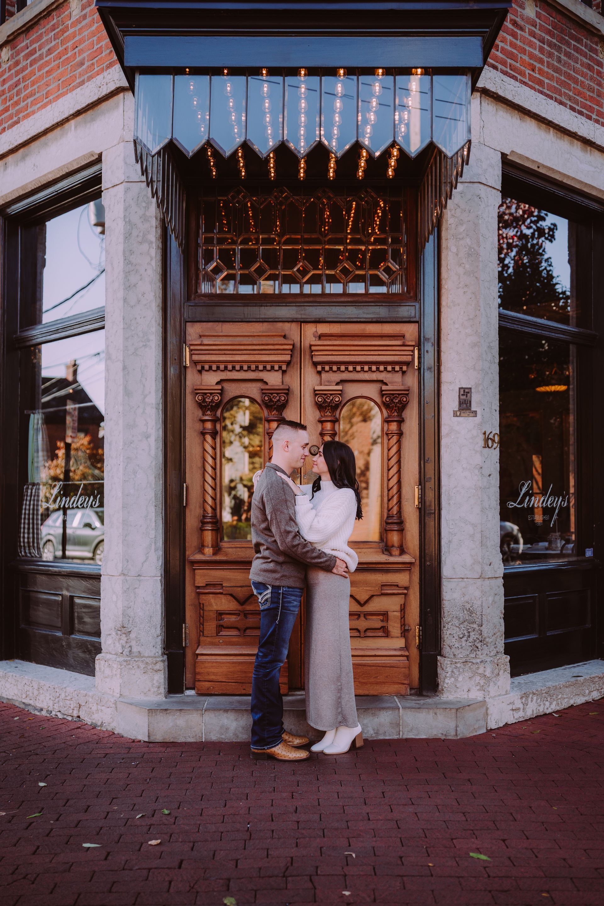 Couple embraces in front of ornate wooden doors of a brick building.