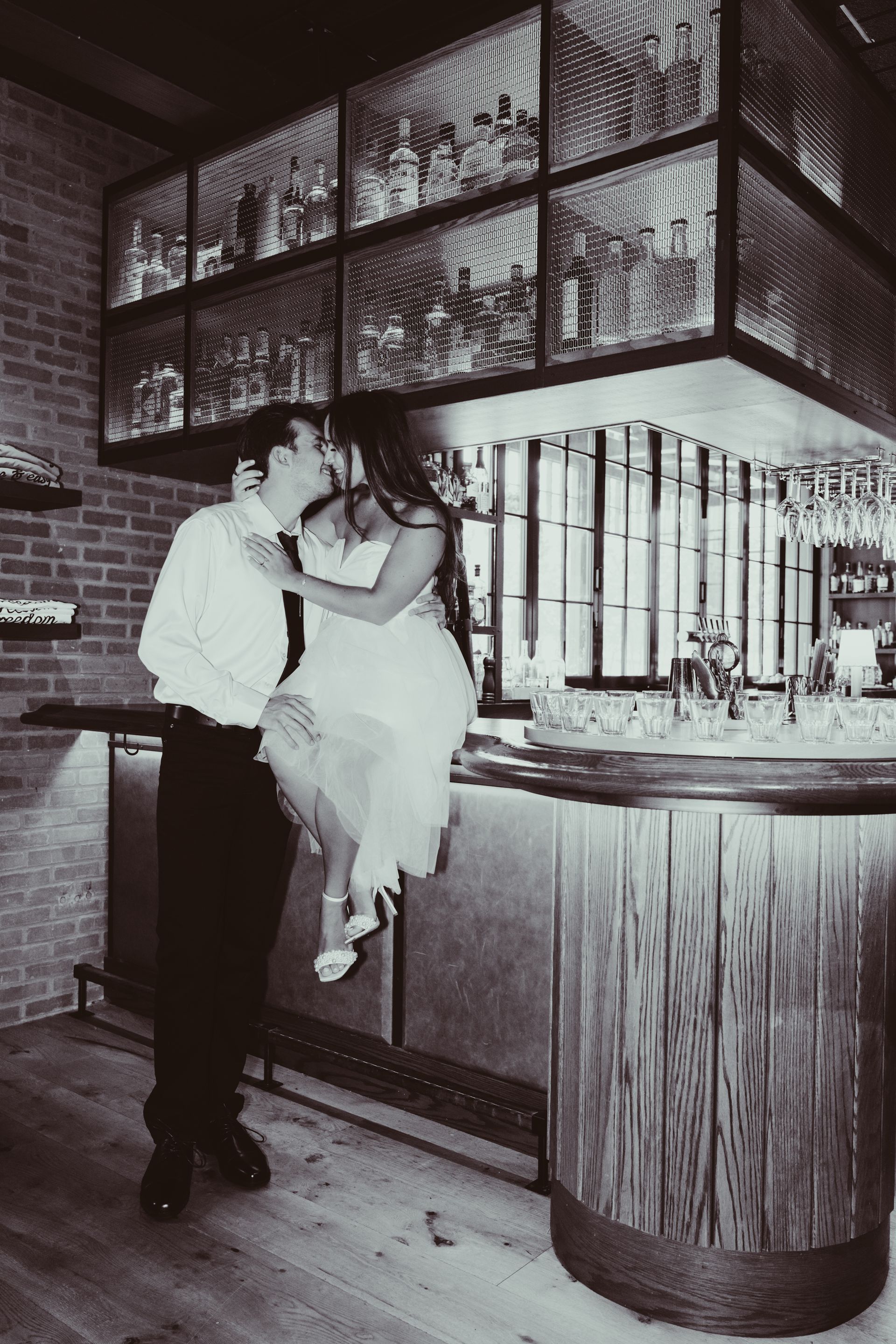 A black and white photo of a man kissing a woman sitting on a bar.